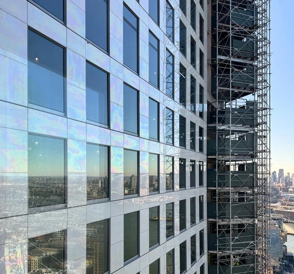 A tall modern glass building with reflective windows and a construction scaffolding on the side, overlooking a cityscape.