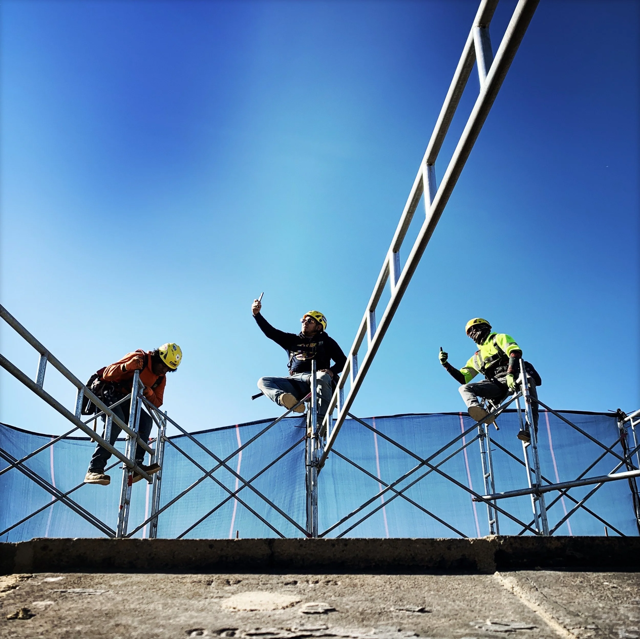 Three construction workers wearing safety helmets sitting on scaffolding, with one taking a selfie, against a bright blue sky.