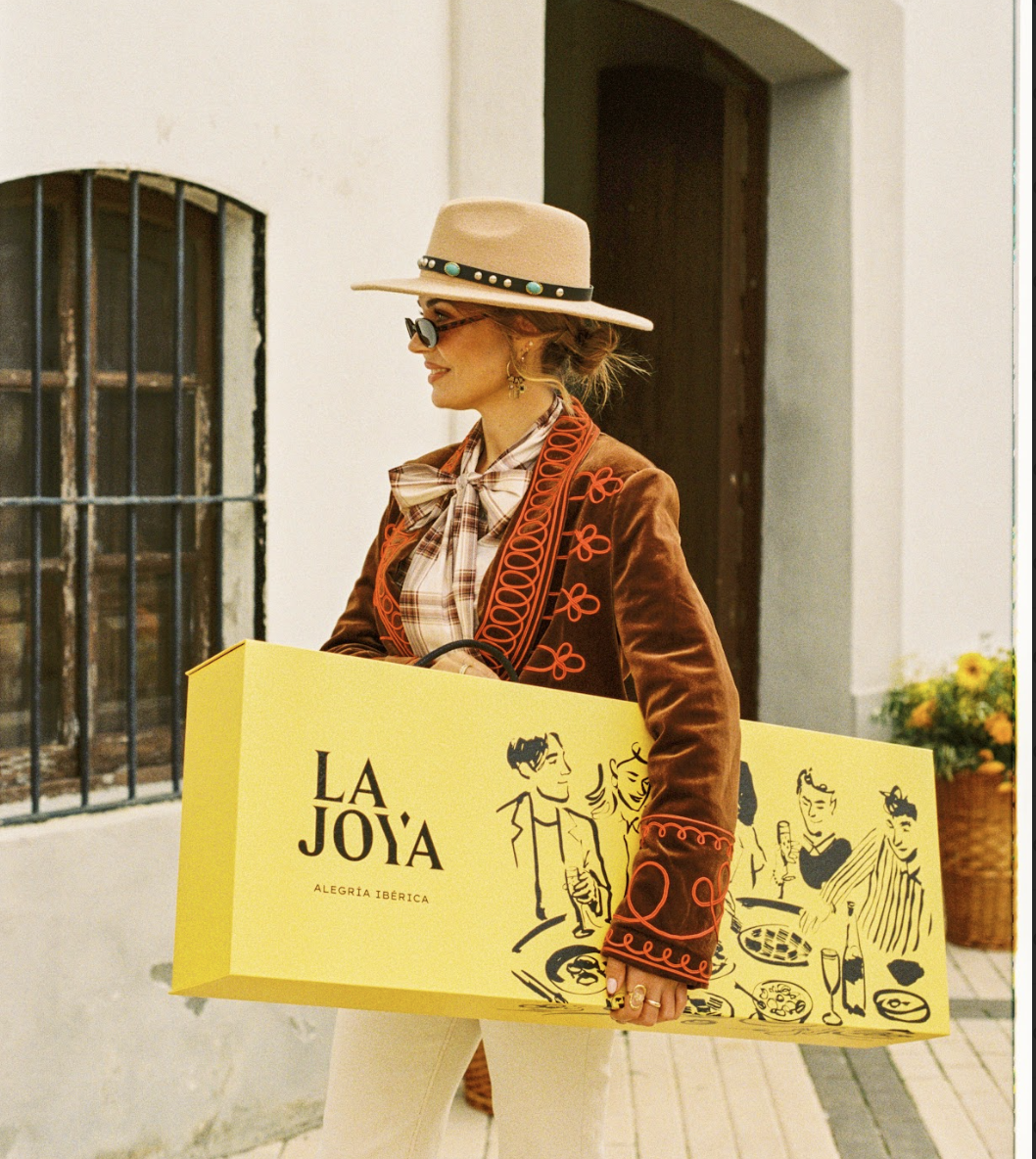 A woman in fashionably vintage attire carrying a large yellow shopping bag with stylized illustrations and the words 'La Joya' and 'Alegría Ibérica'. She wears a beige wide-brimmed hat with a turquoise adornment, sunglasses, earrings, a plaid shirt with a bow tie, and a brown jacket with red embroidered details.