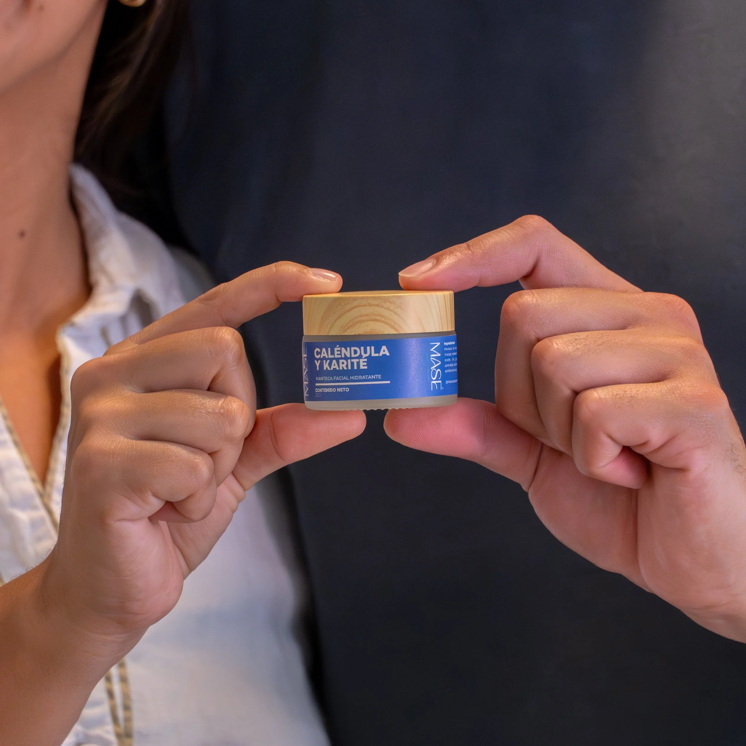 Mano sosteniendo un bote de crema facial etiquetado como 'Caléndula y Karité' con fondo oscuro.