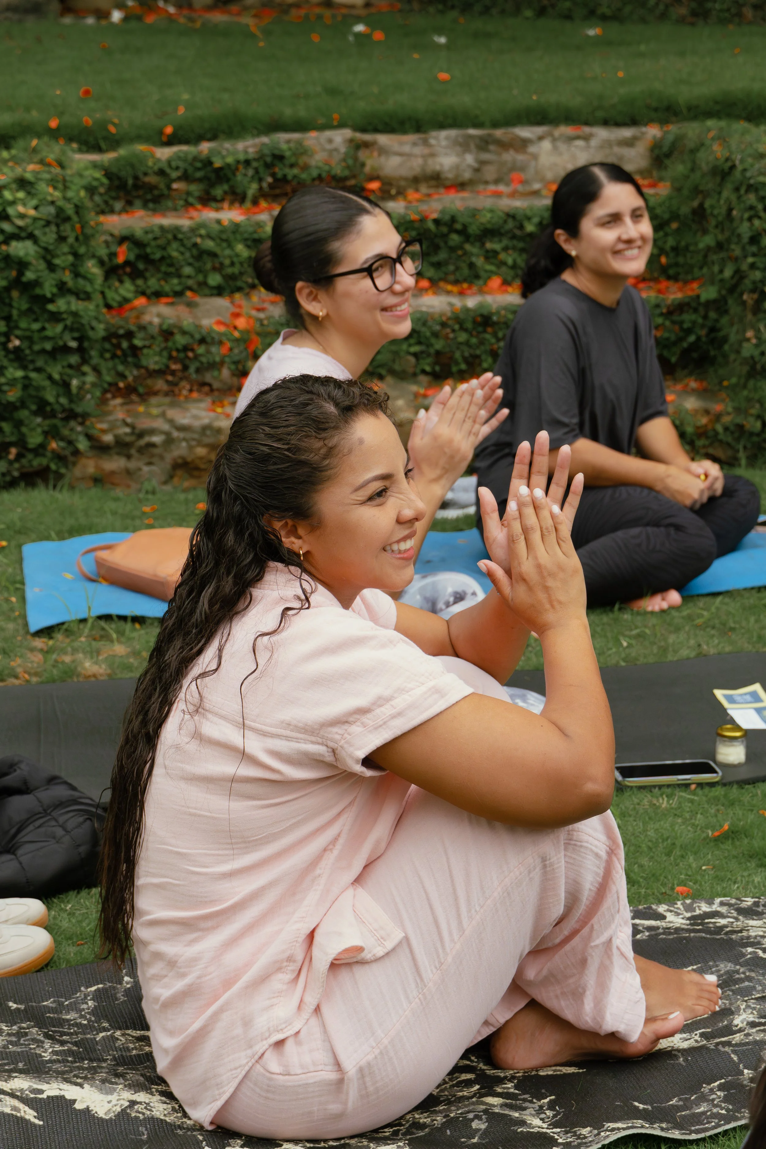 Tres mujeres sentadas en el césped participando en una sesión de yoga o meditación, sonriendo y con las manos juntas en posición de oración, rodeadas de plantas y flores en un jardín.