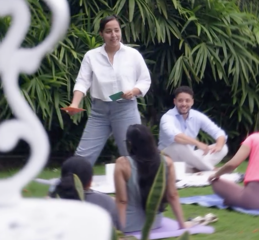 Un grupo de personas participando en una clase de yoga al aire libre rodeadas de plantas verdes. Una mujer en el centro está hablando y todos parecen estar atentos y relajados.