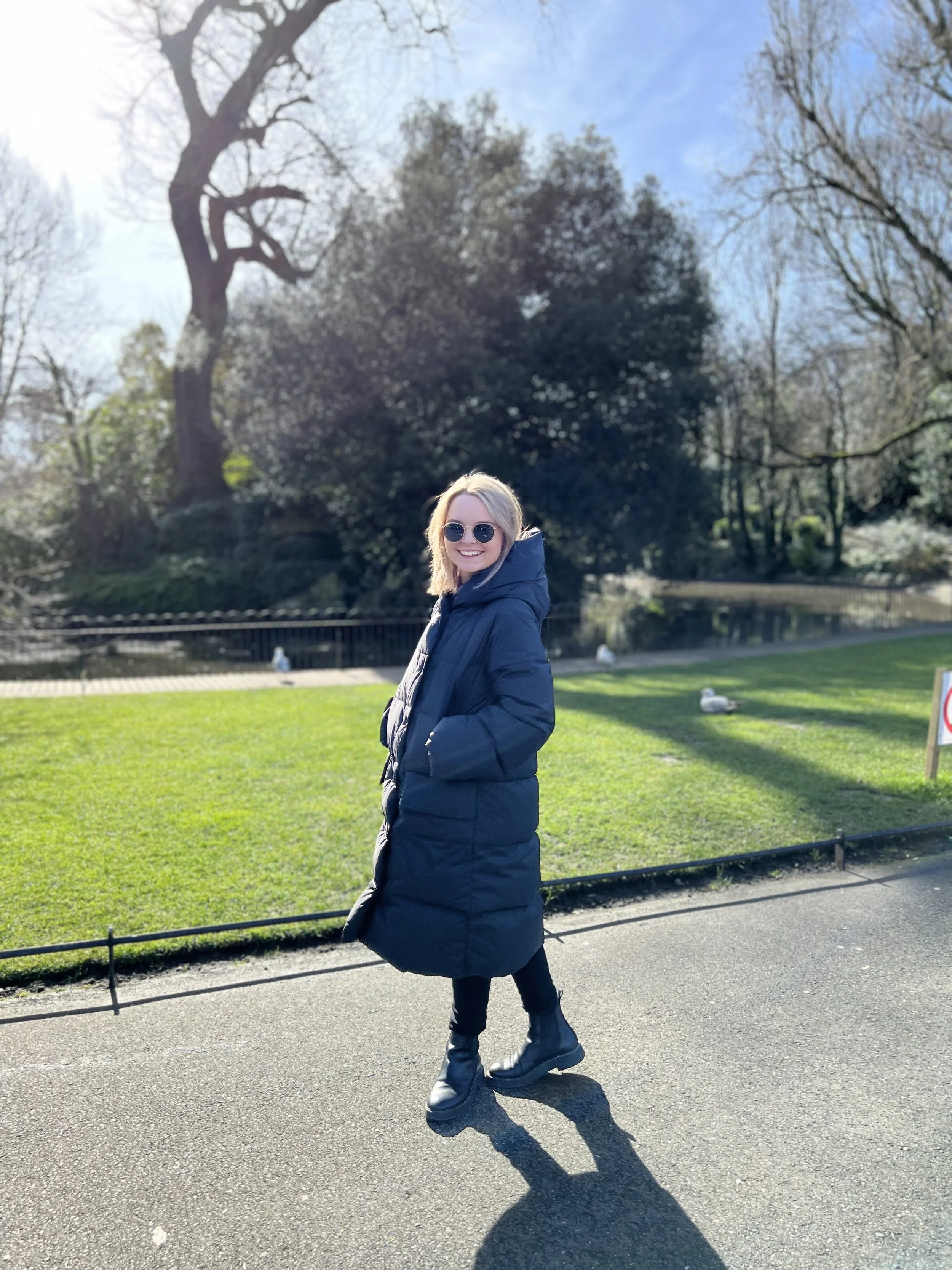 A woman standing on a paved path in a park, wearing a long black coat, black boots, and sunglasses, smiling at the camera with green grass, trees, and water in the background on a sunny day.