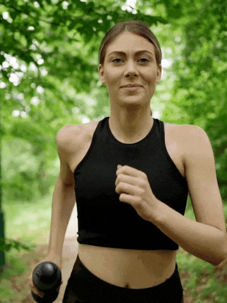 Woman jogging outdoors with a running stance, holding a kettlebell in her right hand, surrounded by green trees.