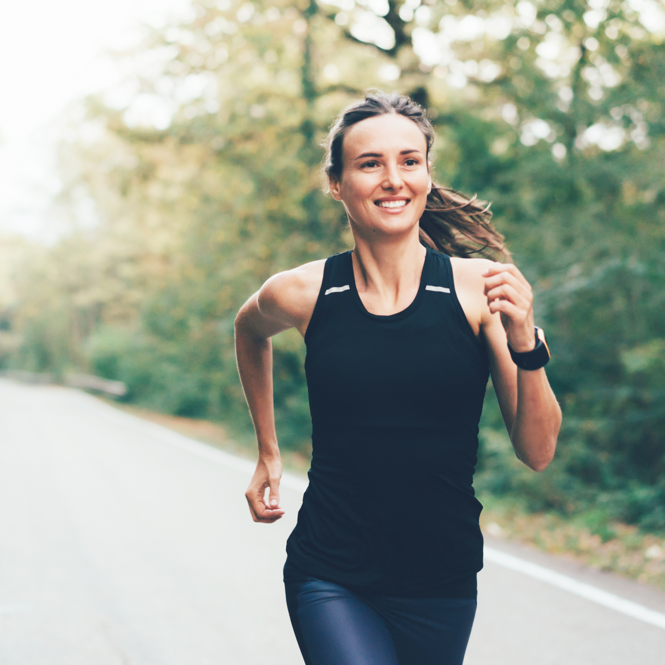 A young woman running outdoors on a tree-lined road, smiling, wearing black athletic clothing and a sports watch.