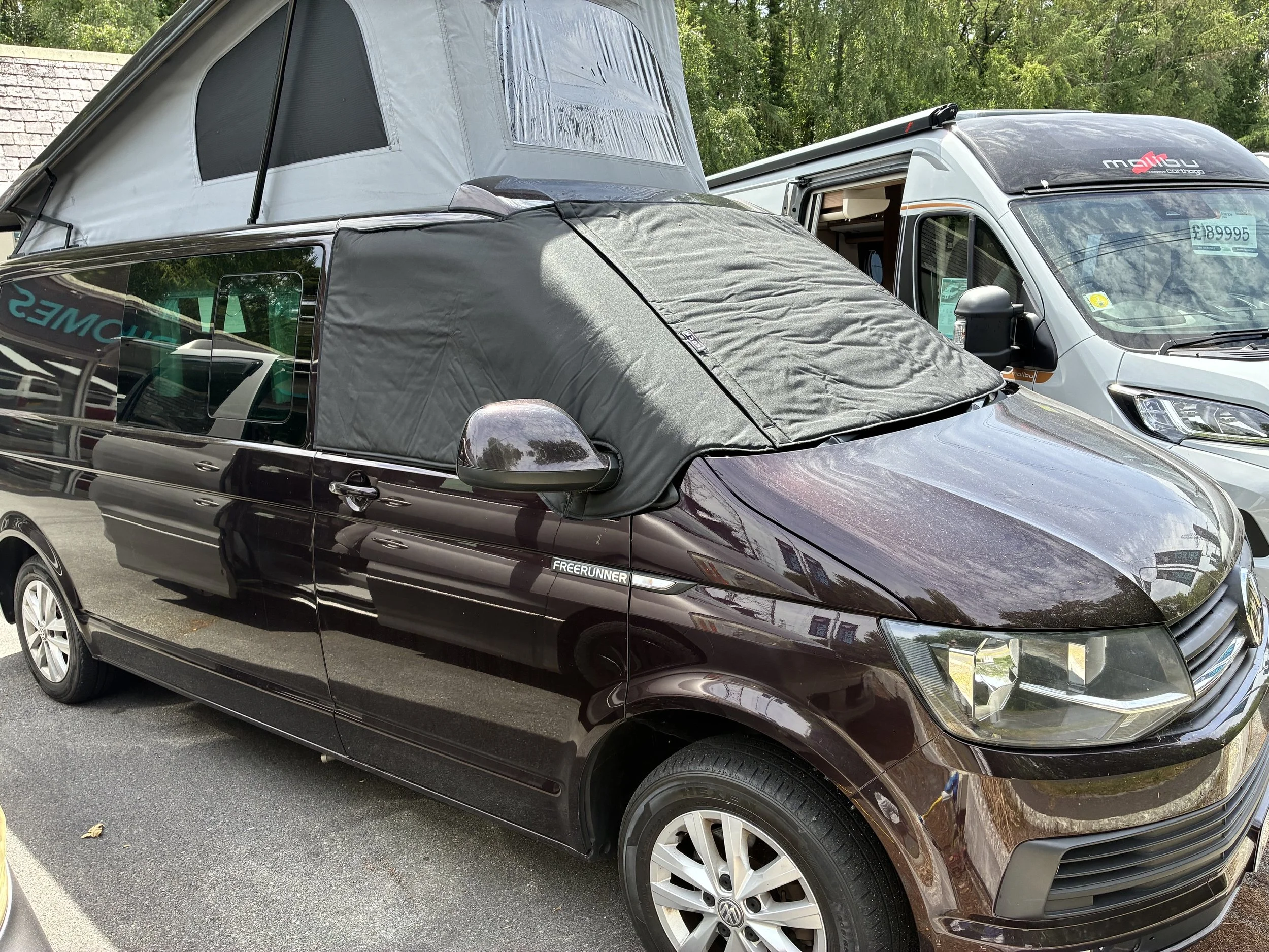 A black Volkswagen Freerunner van with a rooftop tent and a gray window cover parked in a parking lot next to a white camper van with a partial name visible.