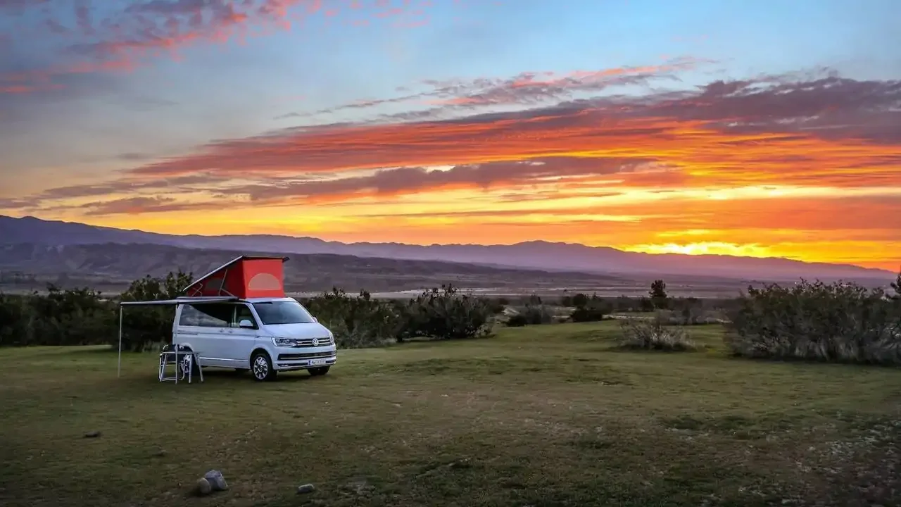 A white van with a red rooftop tent set up on a grassy field at sunset, with mountains and a colorful sky in the background.