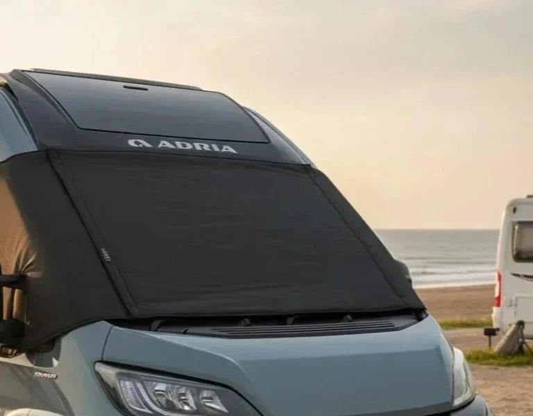 An Adria van parked near a beach with a black sunshade on the windshield. In the background, the ocean and a cloudy sky are visible.