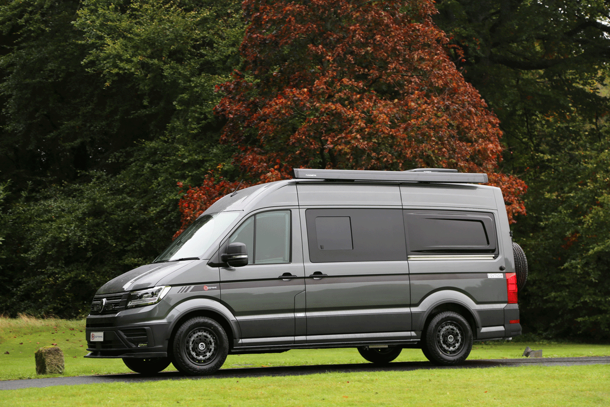 Gray camper van parked on a grassy area with trees showing autumn foliage in the background.