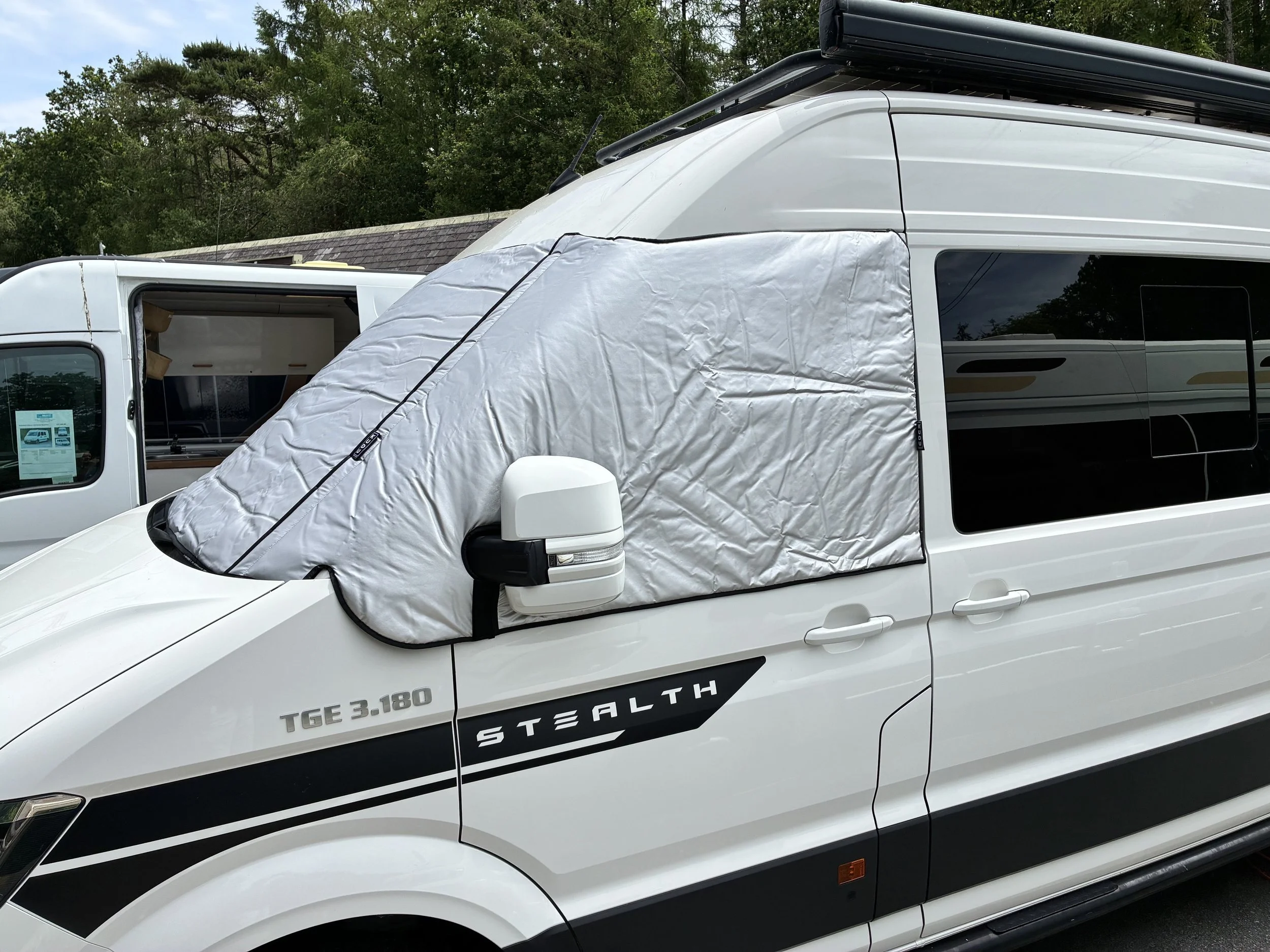 White STEALTH RV with a large tarp or cover attached to the front, parked outdoors with green trees and a building with a shingled roof in the background.