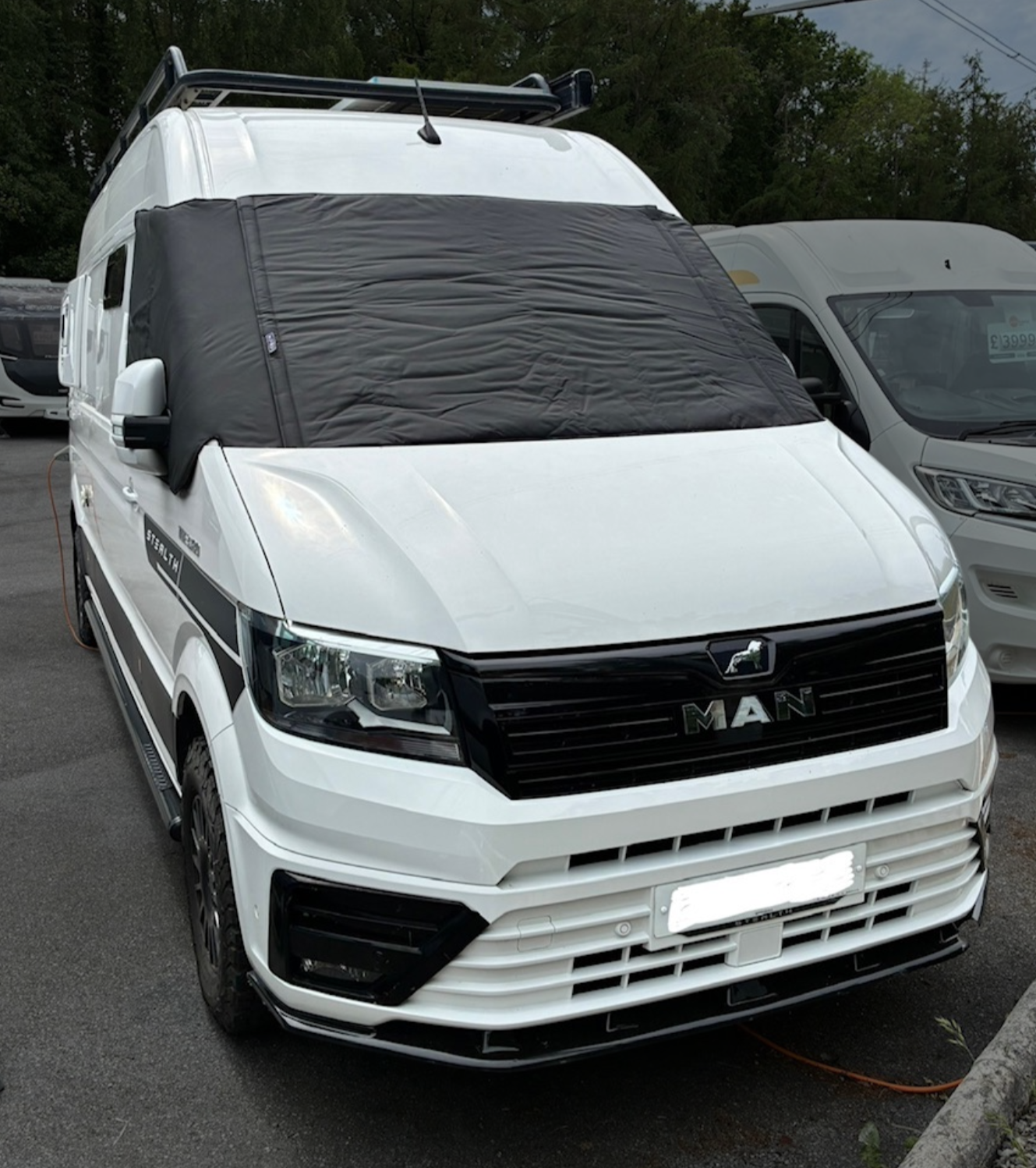 A white MAN van with a black front grille and a black logo, parked outdoors on asphalt, with a large black protective cover over the windshield and part of the side window, and trees in the background.