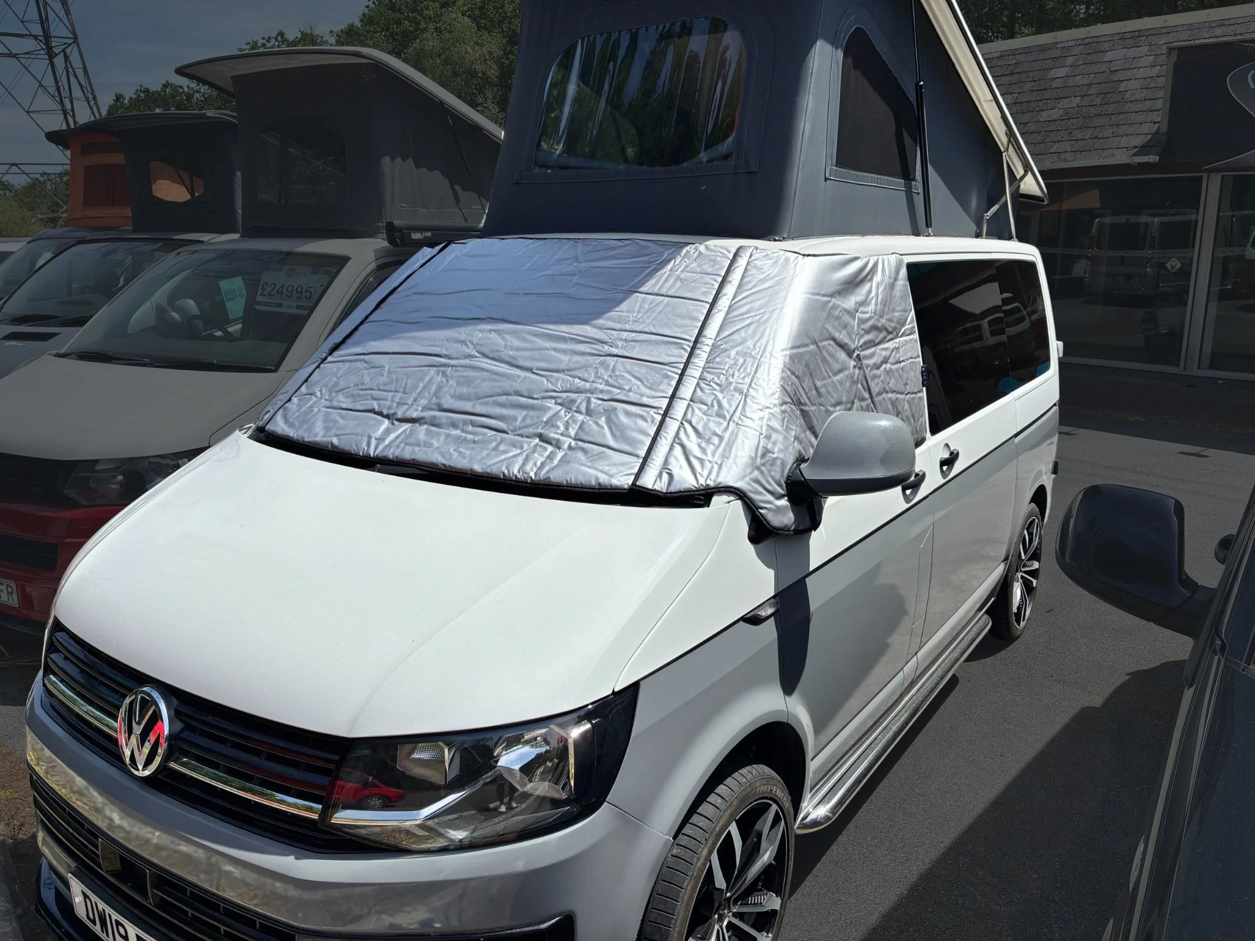 A white Volkswagen van with a gray pop-up roof attached, parked among other cars outside a building with a glass storefront.