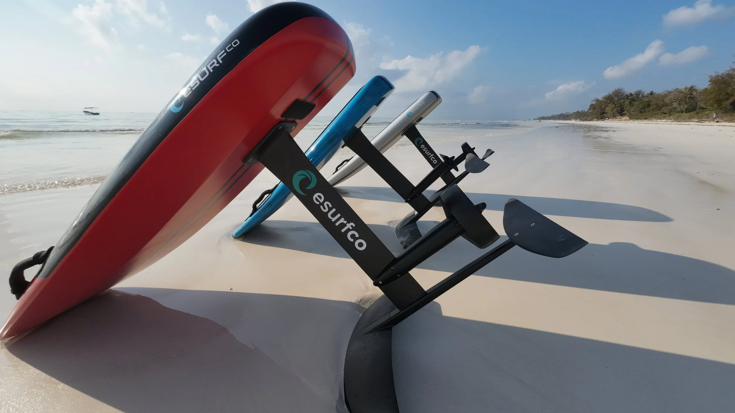Three paddleboards and a rowing machine resting on wet sandy beach near the shoreline with calm ocean waves and distant trees under blue sky with clouds.