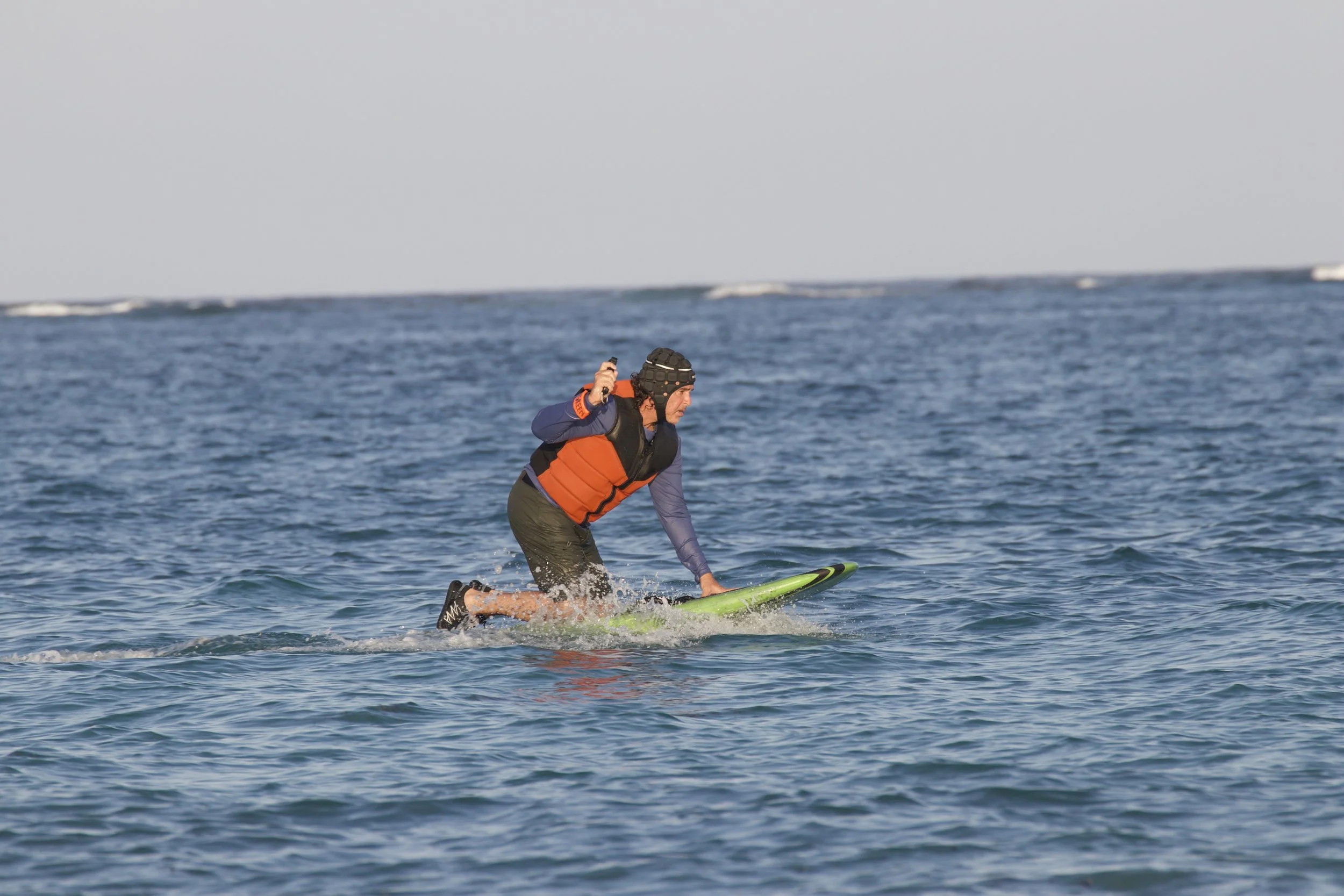 A person wearing a helmet, orange life vest, and black shorts riding a green paddleboard on the ocean with small waves.