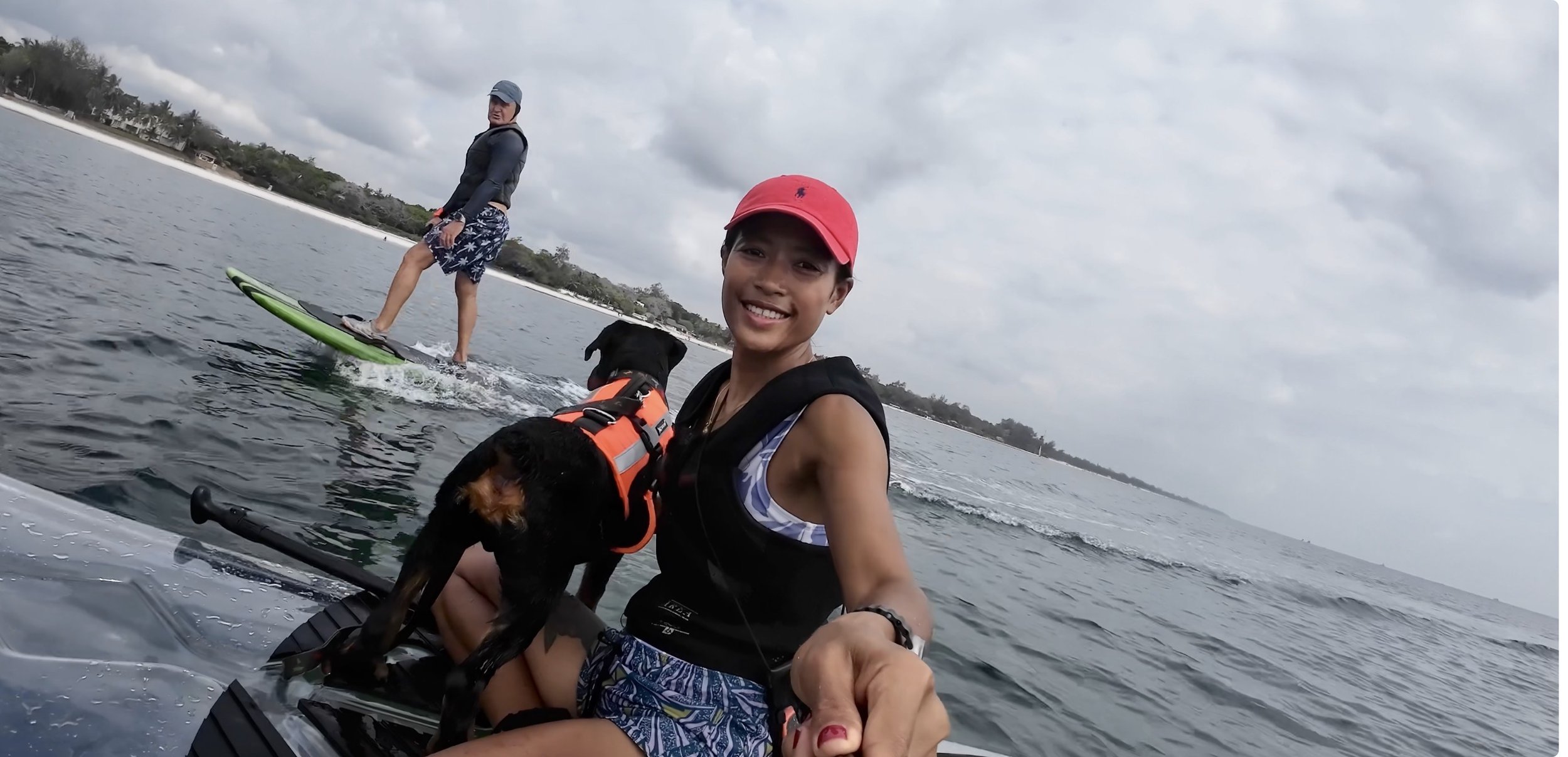 Woman smiling on a boat holding a black dog wearing an orange life jacket, with a man on a paddleboard in the background on cloudy day.