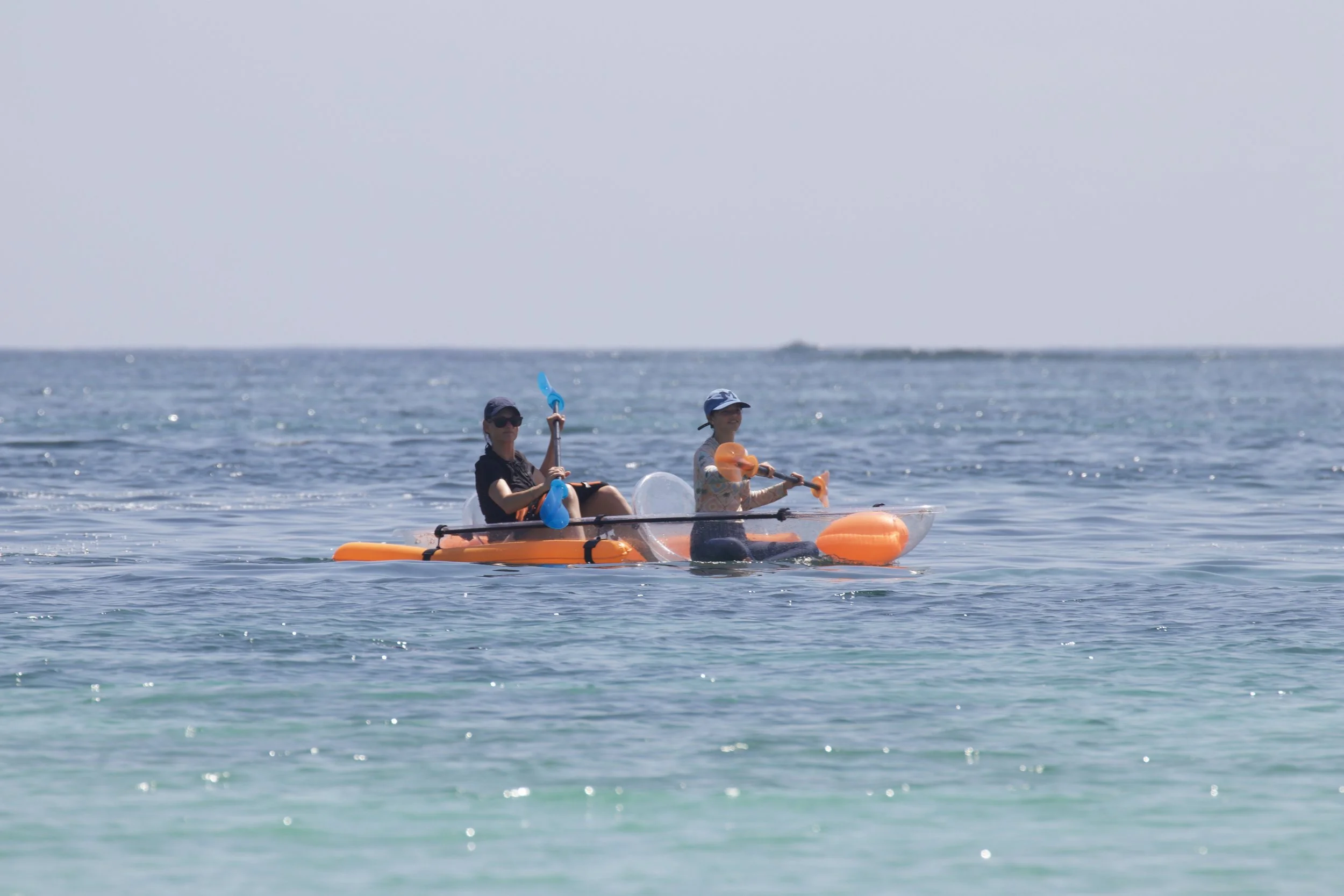 Two people kayaking in the ocean, both wearing hats and sunglasses, in a yellow kayak with orange floatation aids.