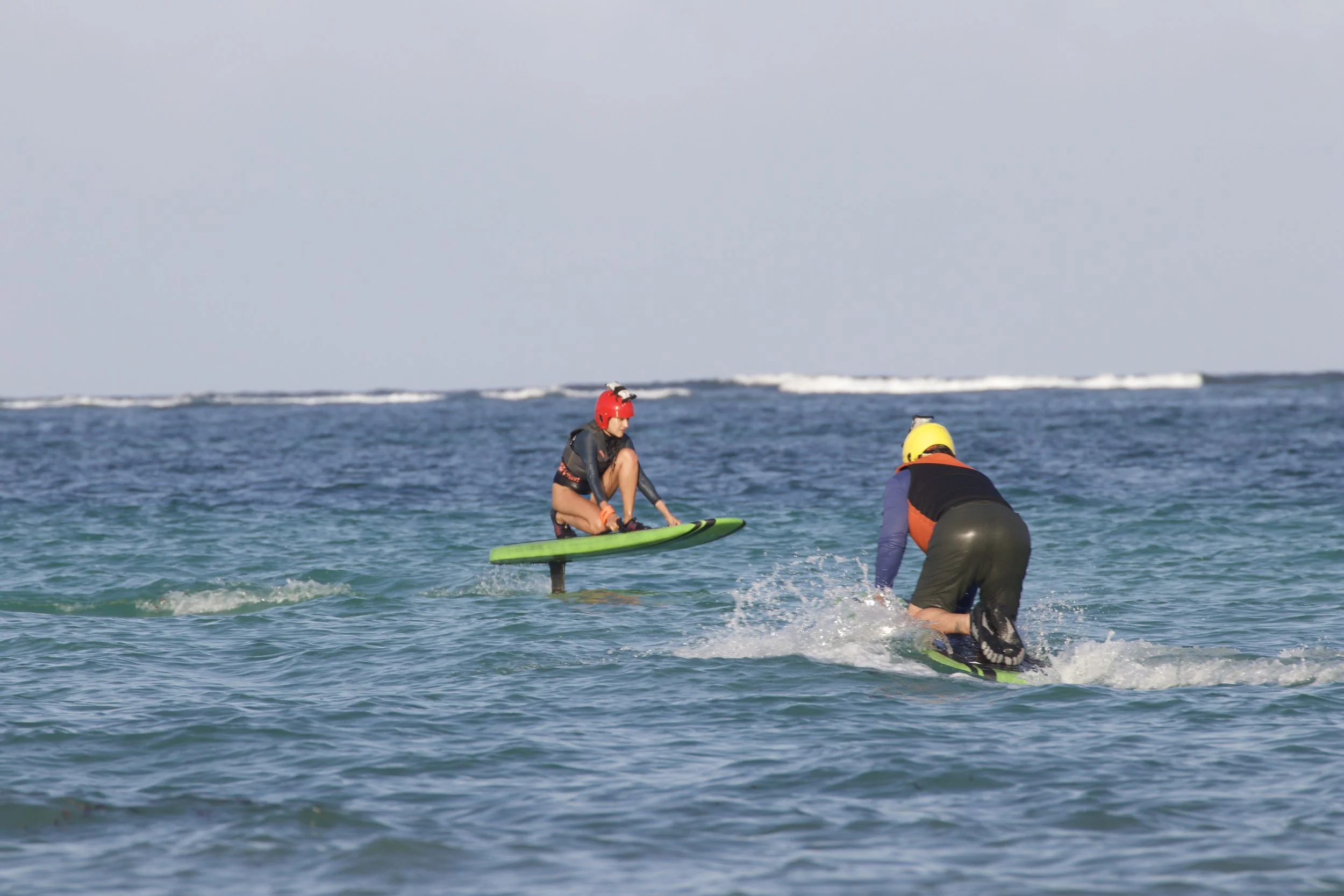 Two people in wetsuits, helmets, and life jackets engaging in water sports; one standing on a paddleboard on the water, the other kneeling on a paddleboard and paddling.
