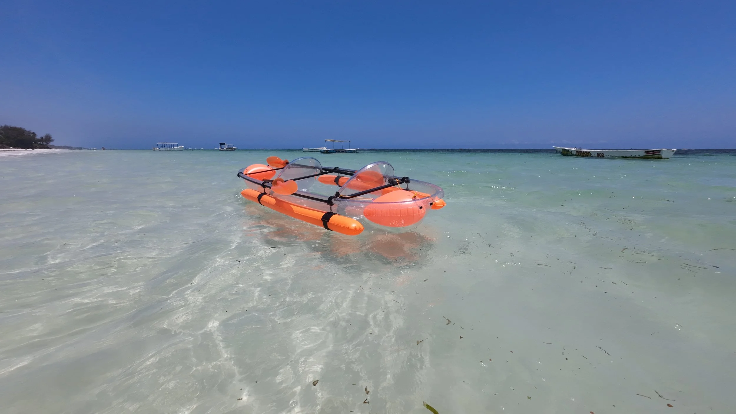 An orange inflatable boat with a clear canopy floating on clear shallow water at the beach with boats in the background under a clear blue sky.
