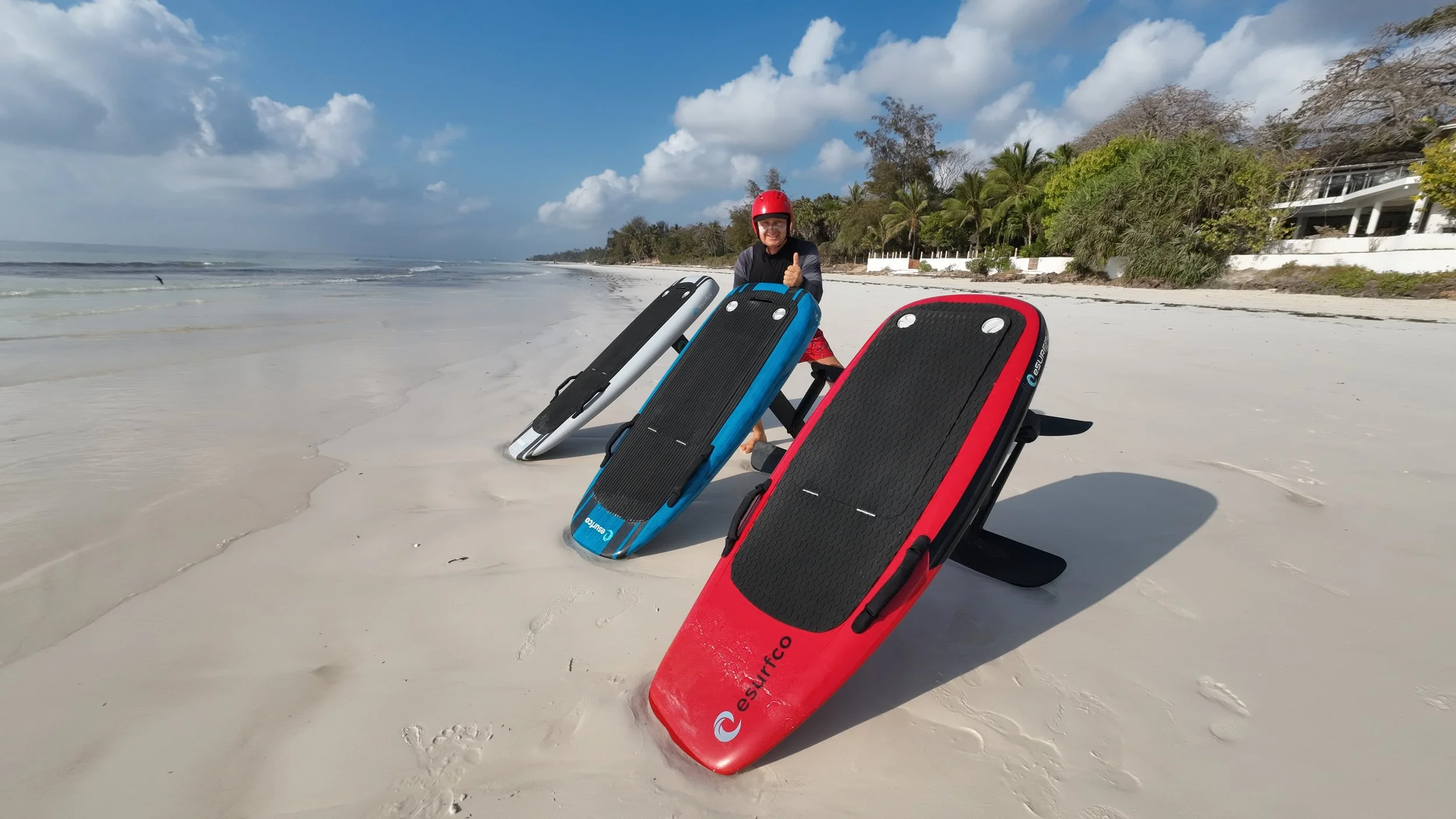 A man wearing a red helmet kneeling on a beach with three colorful paddleboards, giving a thumbs-up gesture.