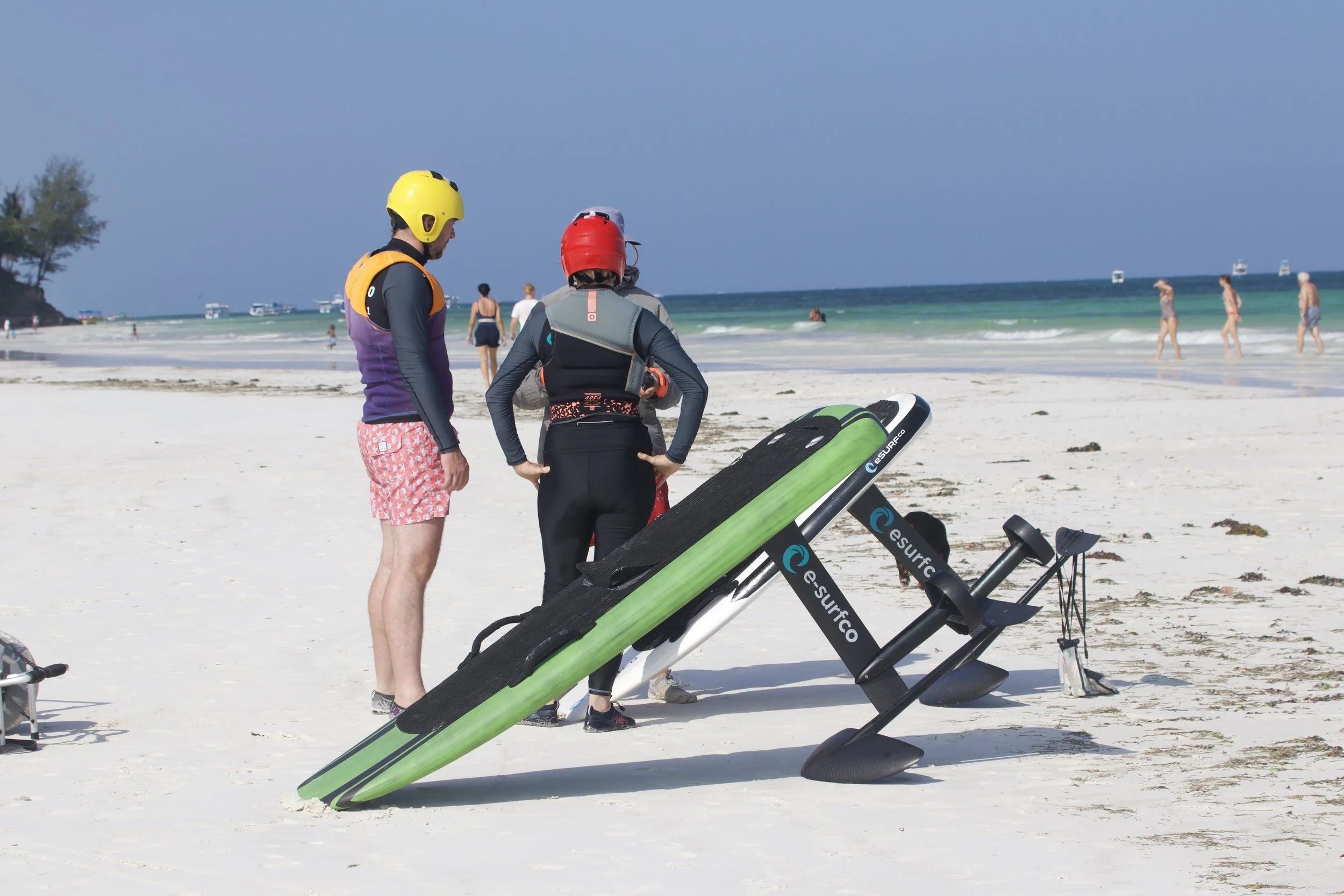 Two people wearing safety helmets and wetsuits standing on a beach near a green motorized surfboard, with others swimming and walking in the background.