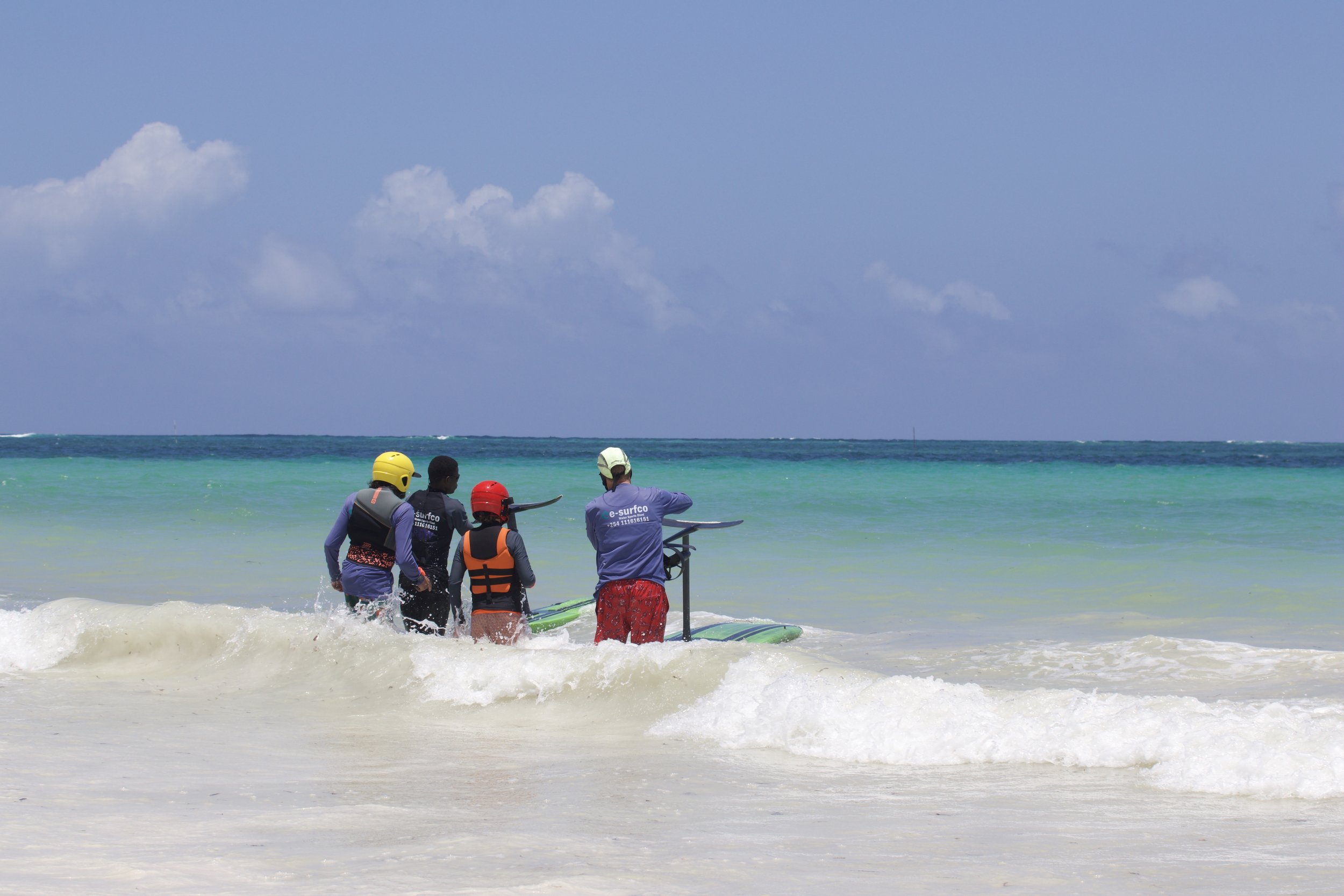 Group of four people preparing to go paddleboarding in the ocean, wearing life jackets and helmets, near a sandy beach with blue sky and turquoise water.
