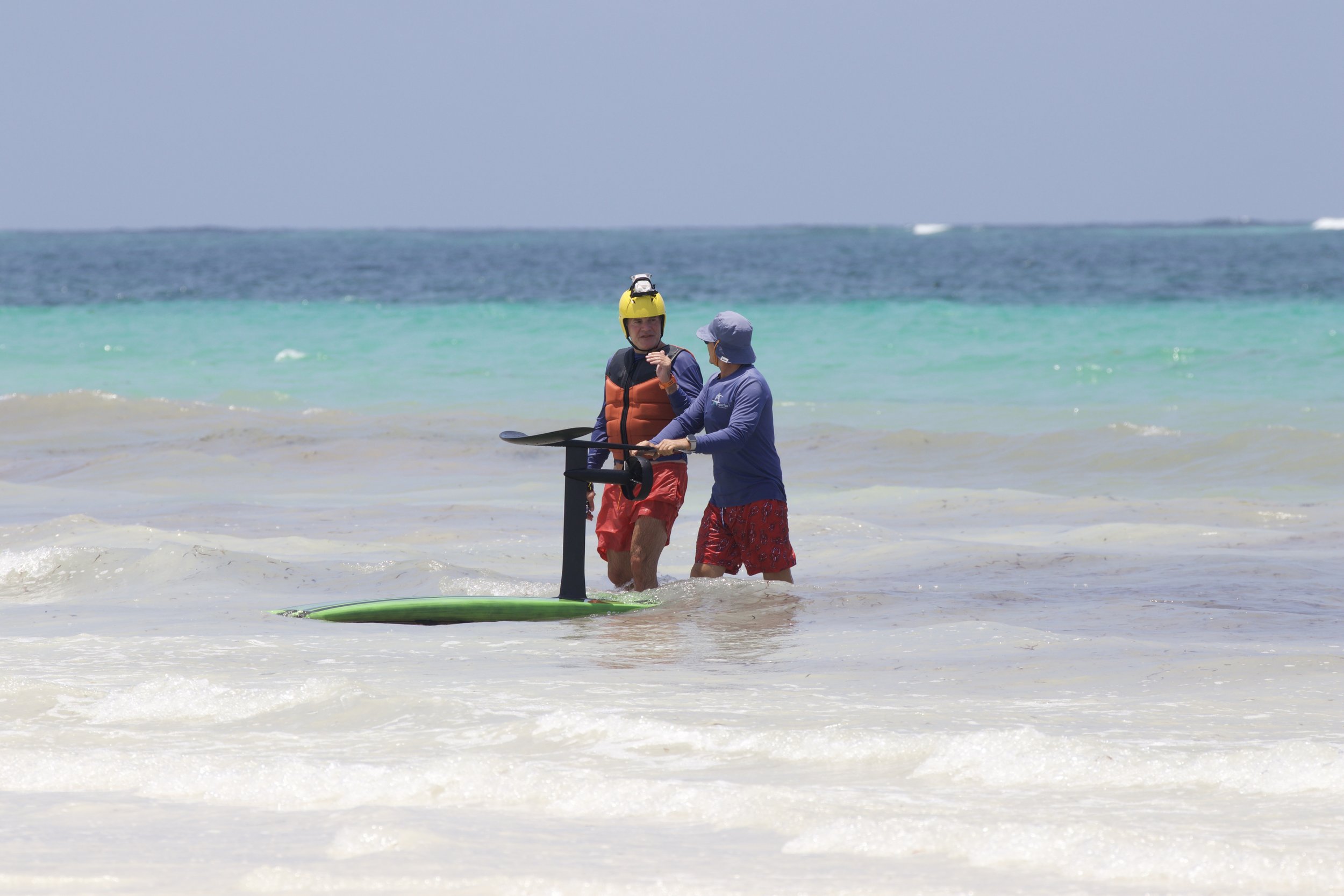 Two people standing in shallow ocean water, one is wearing a helmet and safety gear, the other is holding a paddleboard.