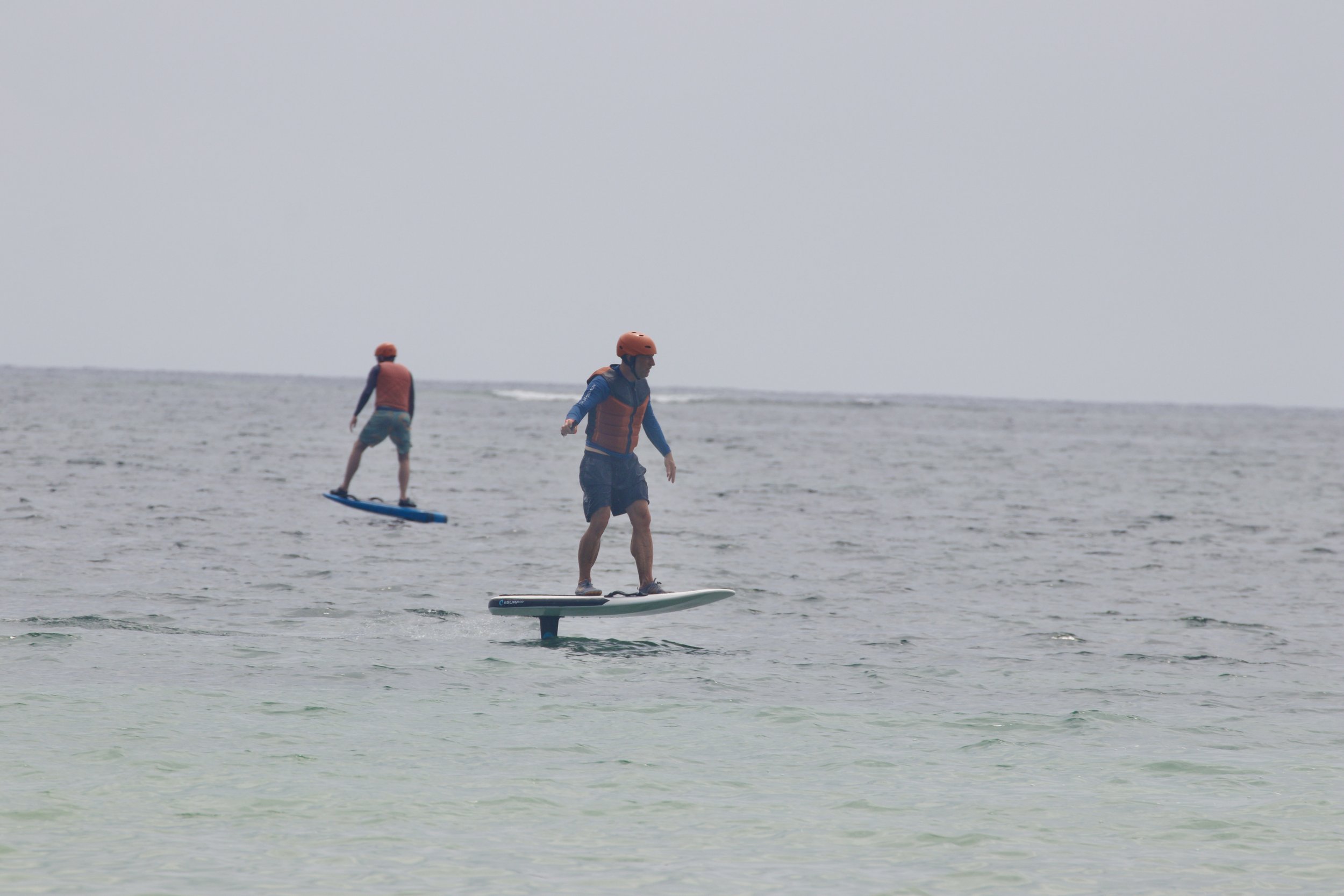 Two people riding electric surfboards on the ocean water, both wearing helmets and life jackets.