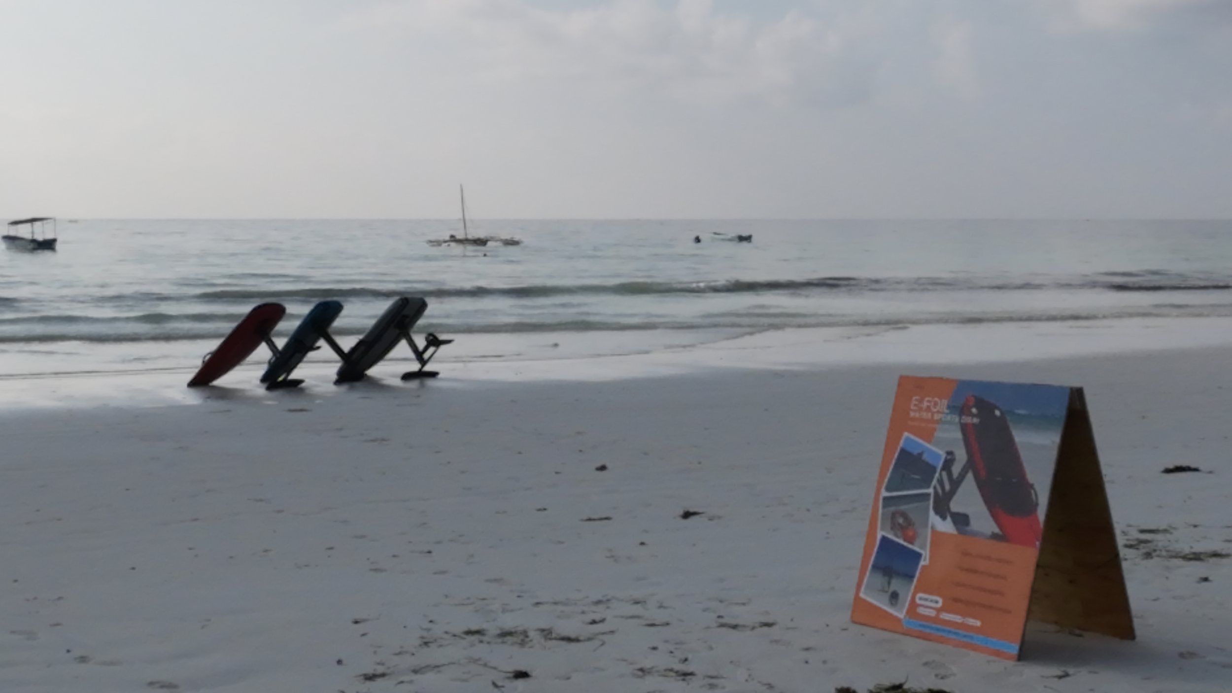 A sandy beach with three folded chairs near the water's edge, palm trees and boats in the distance, and a promotional sign advertising a product on the sand.