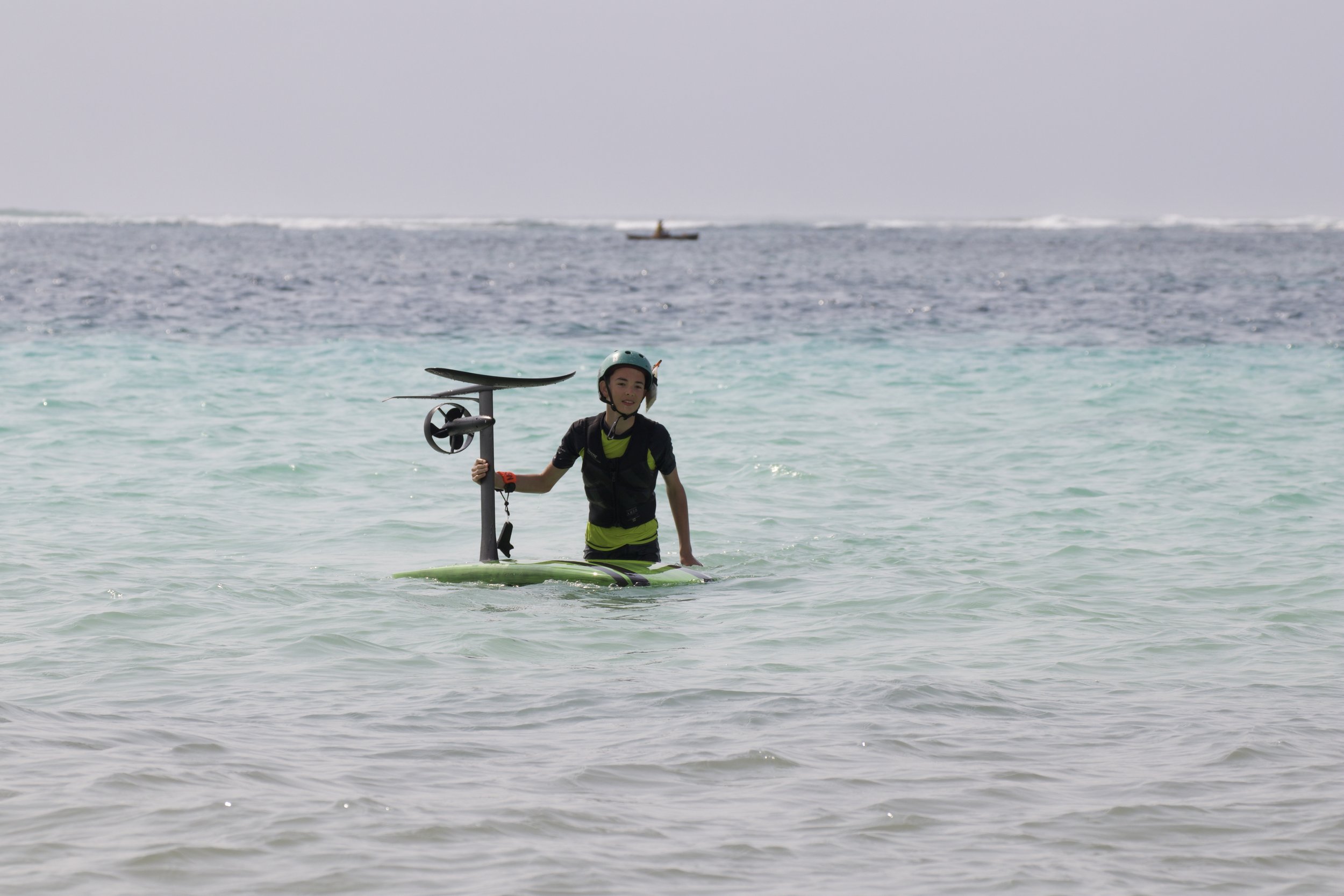 A person standing on a paddleboard in the ocean, holding a sail with a windsurfing sail attached, wearing a helmet and wetsuit.