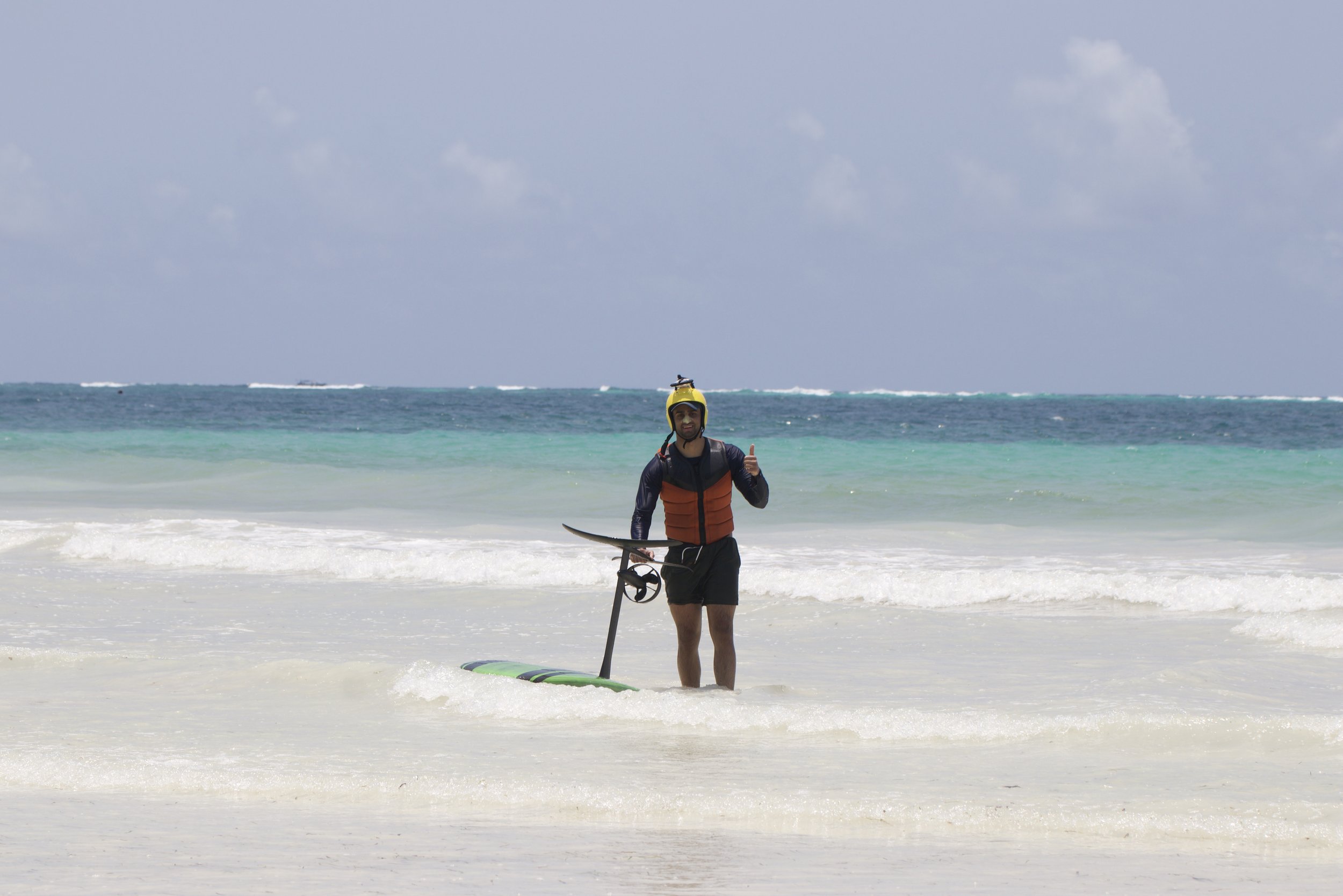 Man in wetsuit and life jacket standing in shallow water on the beach, holding a paddleboard and giving a thumbs up, with ocean and cloudy sky in the background.