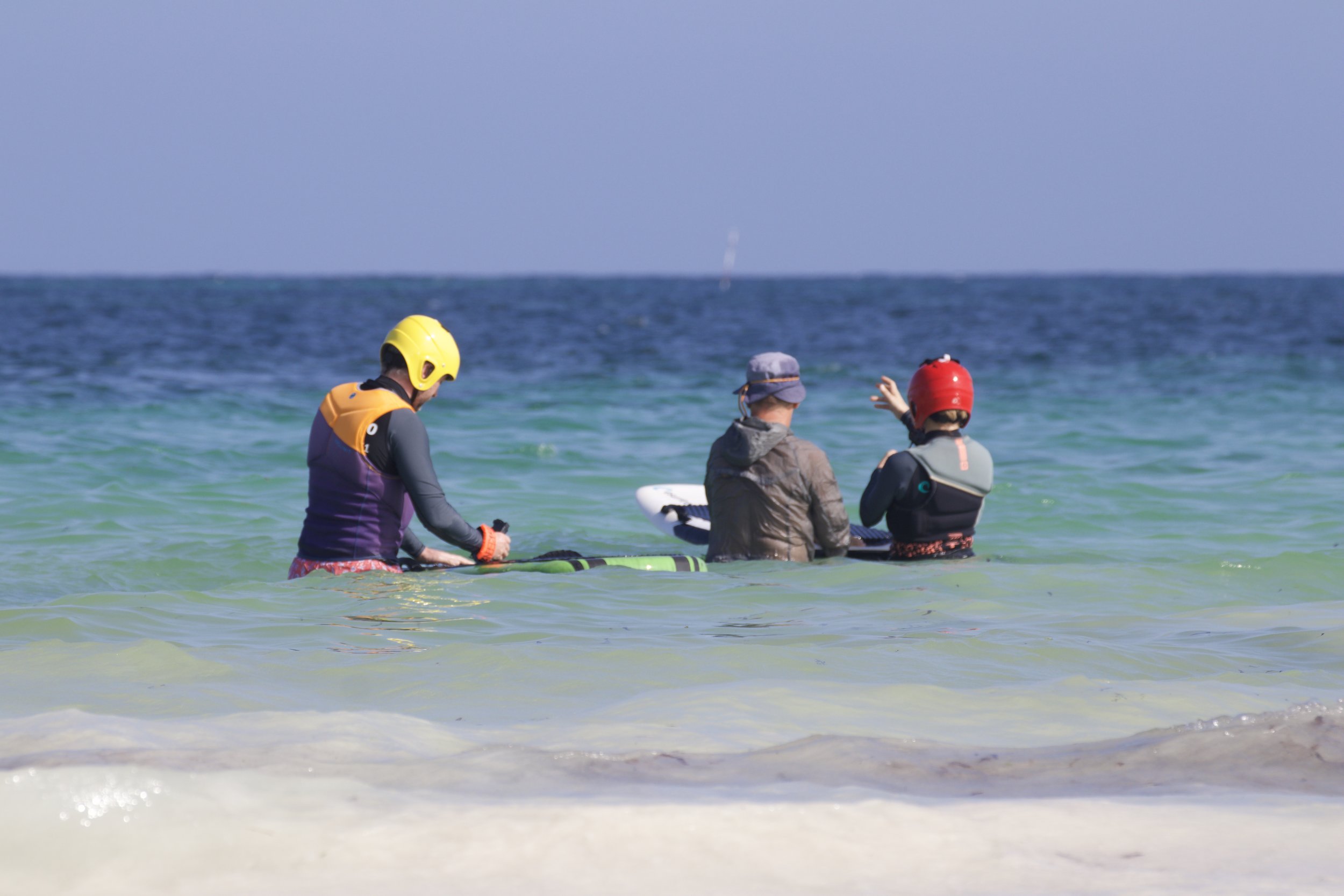 Three people wearing helmets and life jackets standing in the shallow water at the beach, with ocean waves behind them.