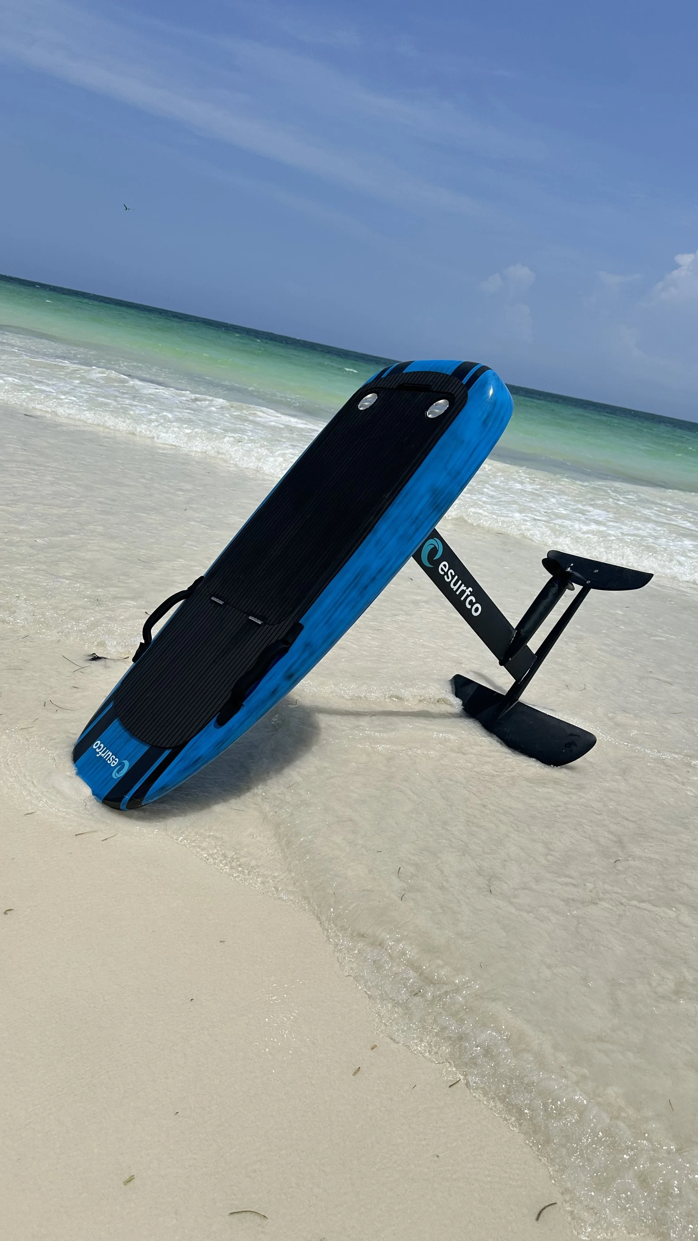 An eSurfco electric surfboard on a sandy beach with gentle waves and a blue sky.