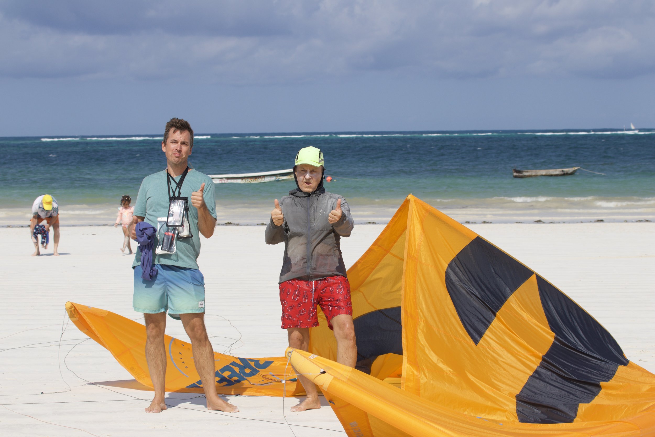 Two men stand on a sandy beach next to a deflated orange and black kite, giving thumbs up. In the background, children and two boats are visible on the water, with a cloudy sky overhead.