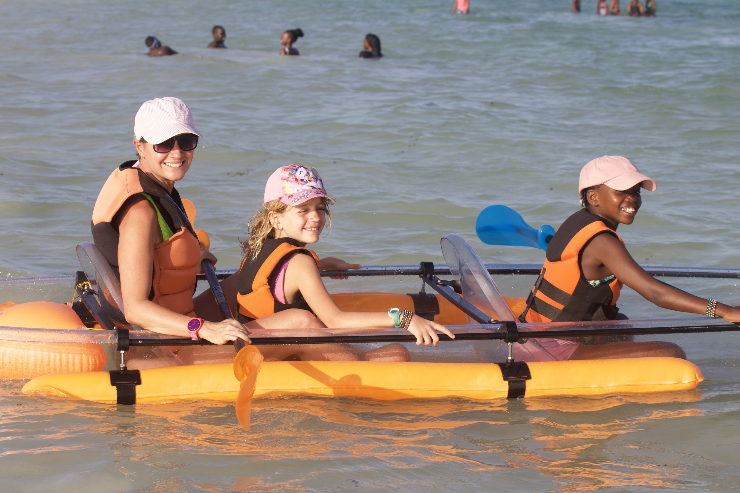 Three people on an orange kayak in the water, two young girls and an adult woman, all wearing life jackets and hats, with other swimmers visible in the background.
