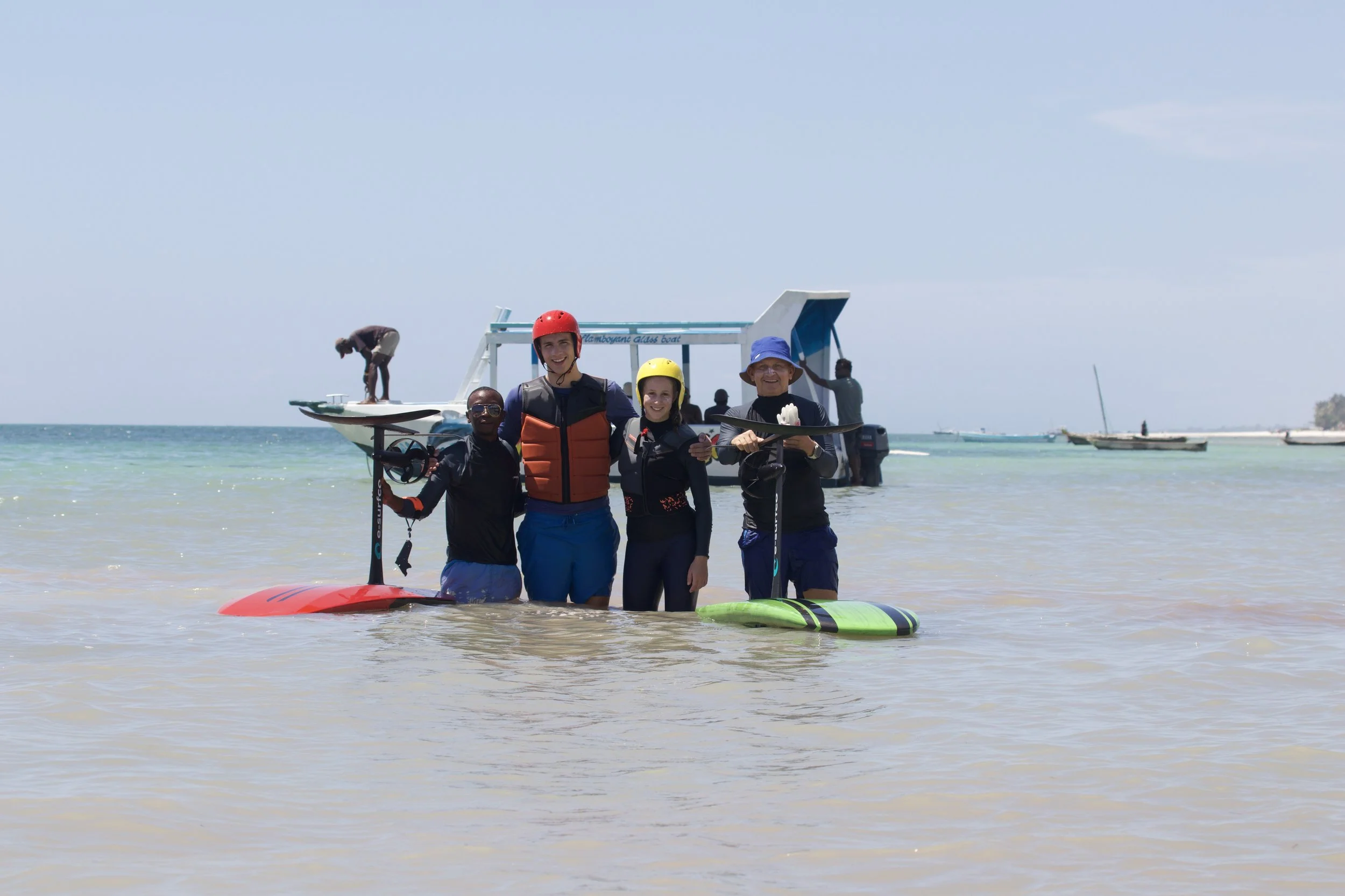 Four people standing in shallow water at the beach, holding paddleboards and windsurfing equipment, with a boat and another person in the background on the water.