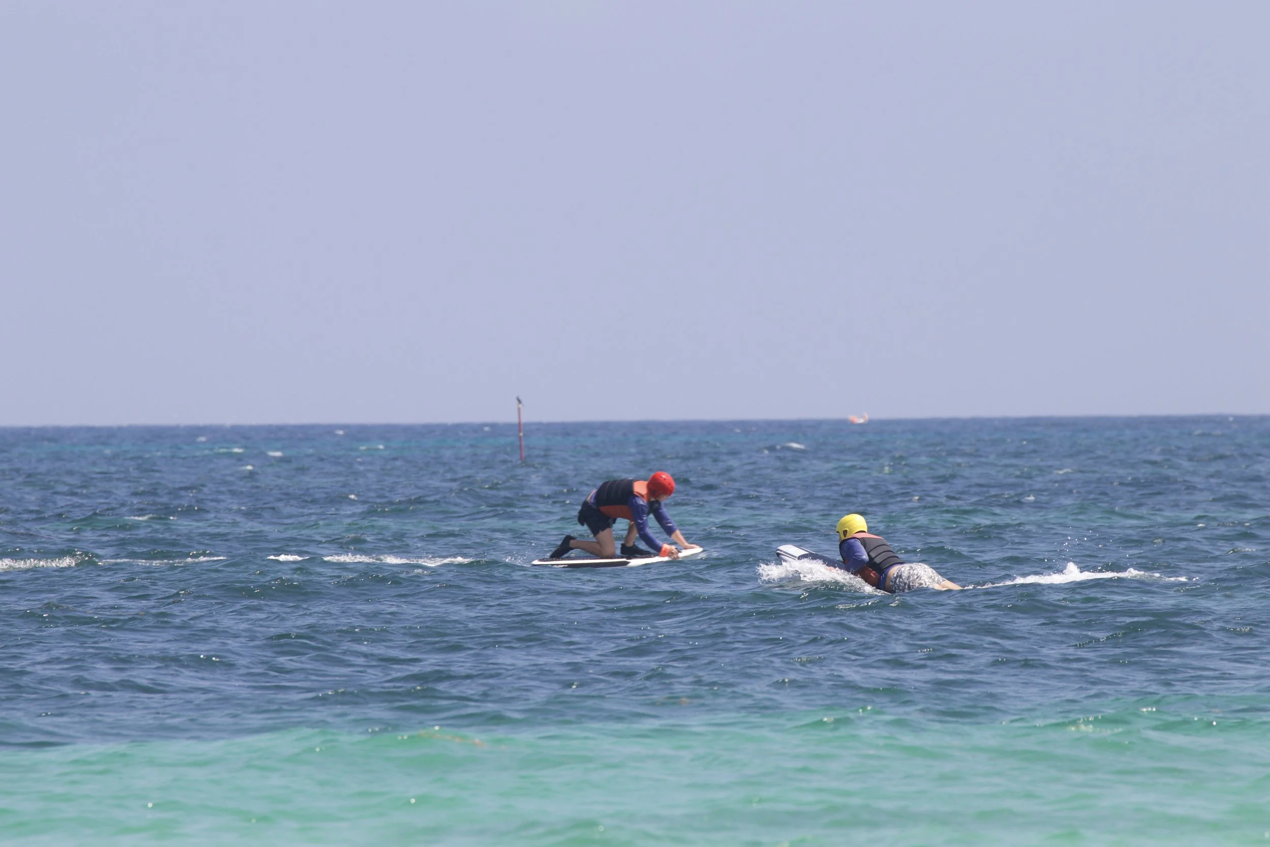 Two people engaging in water sports on the ocean, one kneeling on a paddleboard and another swimming nearby with a yellow helmet, both wearing life jackets.