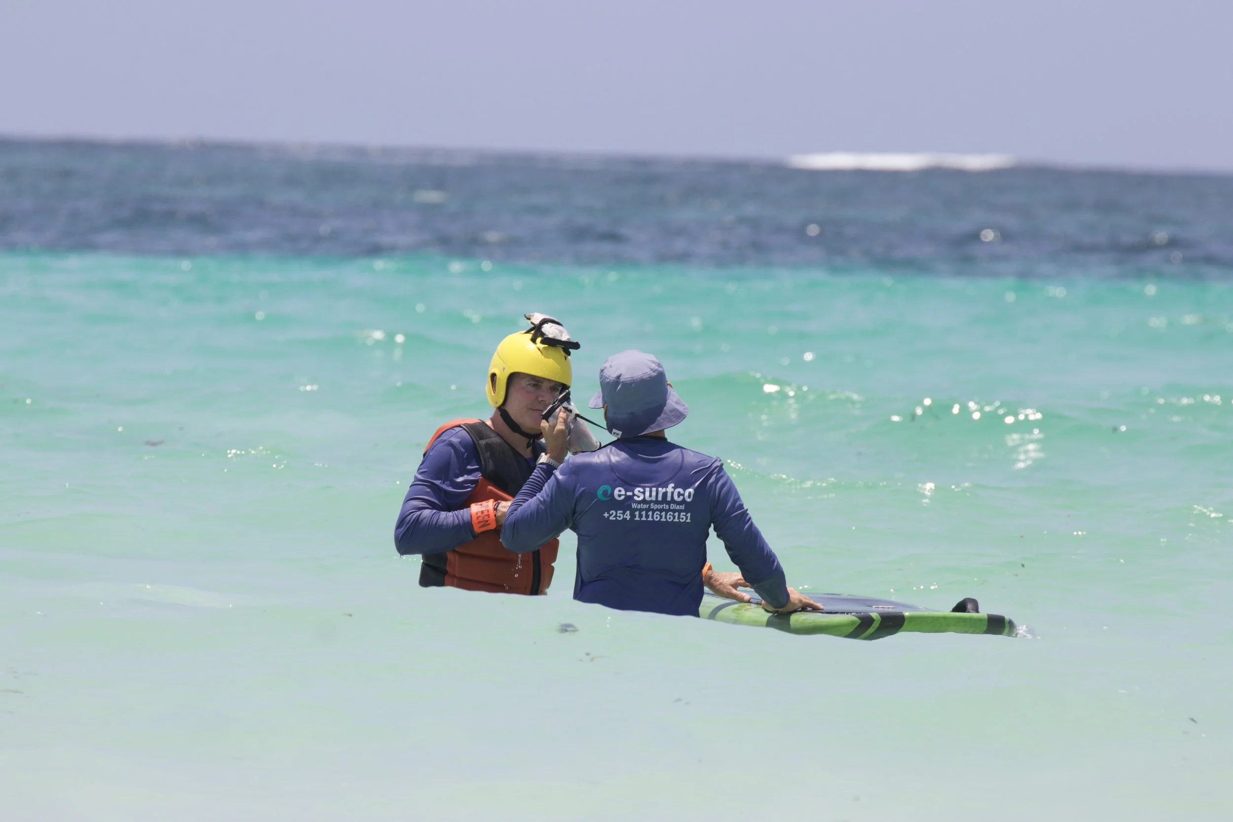 Two people in the water, one wearing a yellow helmet and the other wearing a blue hat and jacket, holding a paddleboard.