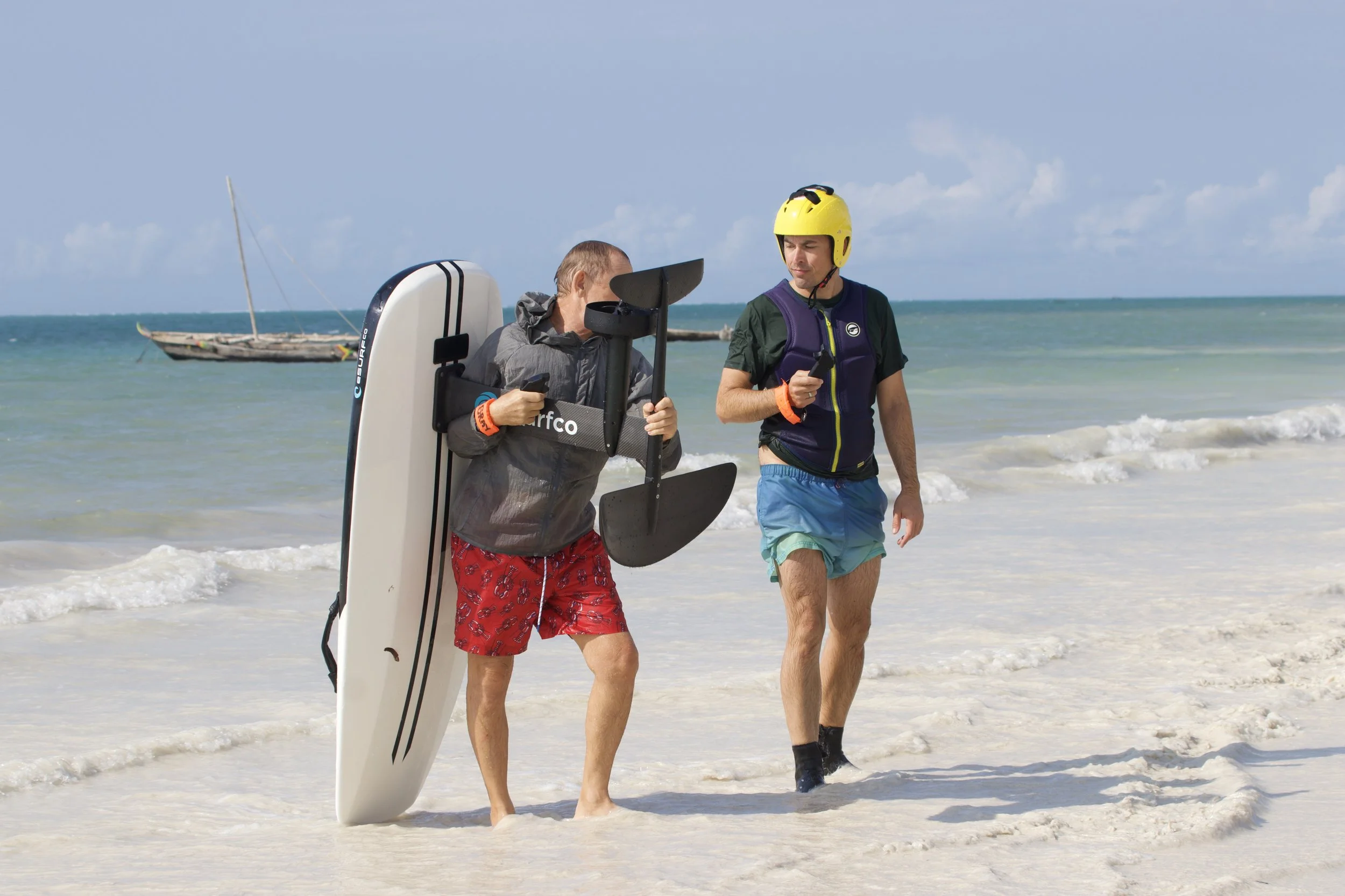 Two men walking on the beach, one holding a surfboard and a shovel, the other with a helmet and a device in his hand, with a boat in the background.