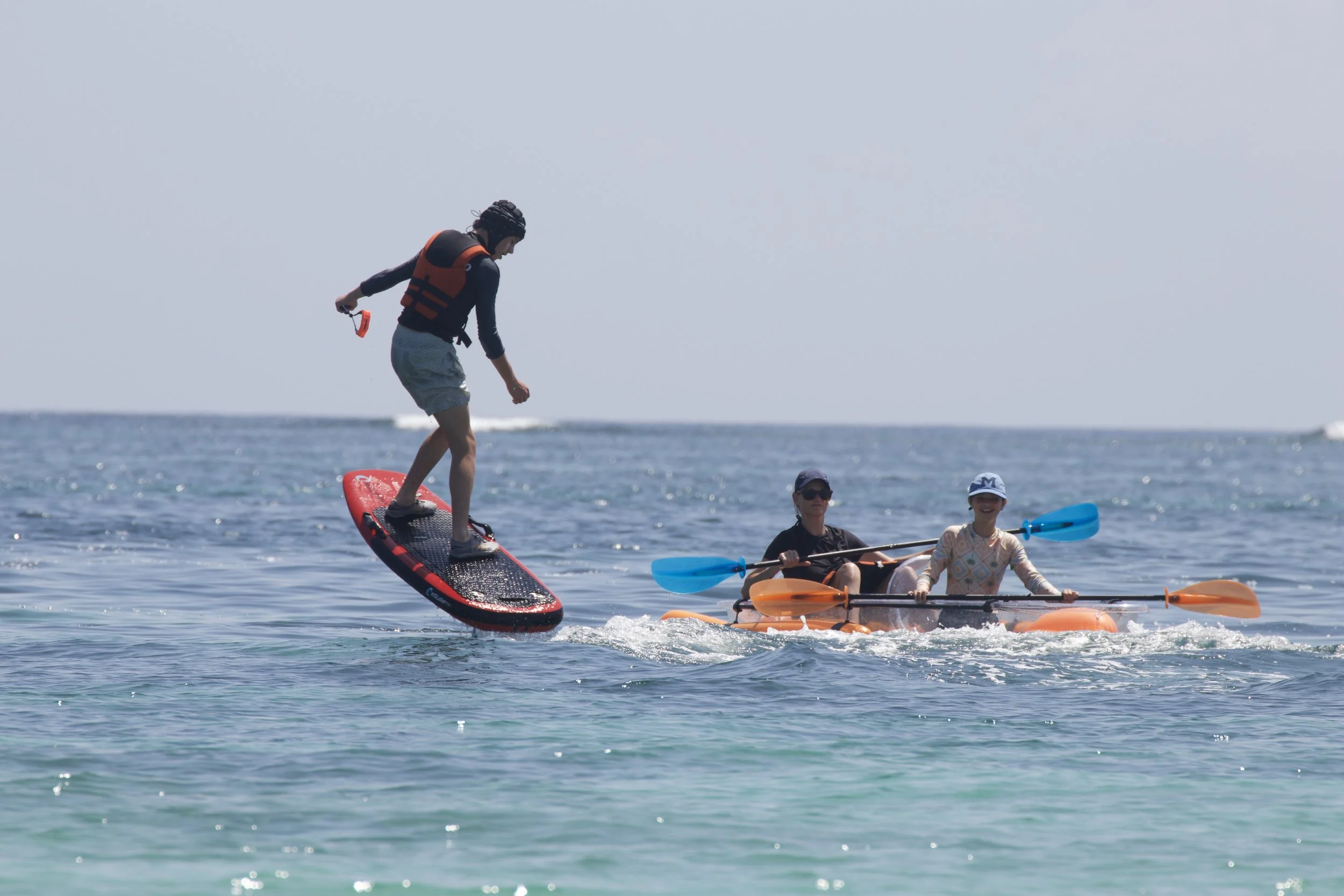 A person is riding a stand-up paddleboard over the water while two others are sitting in an orange kayak with blue and orange paddles on a sunny day at the beach.