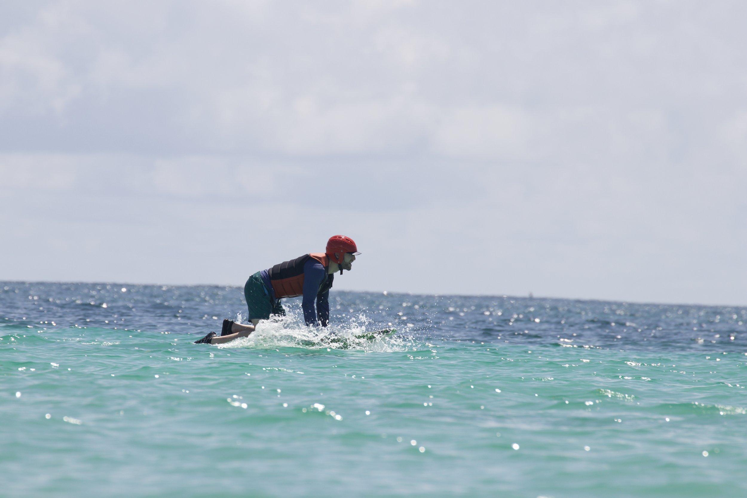 Person wearing a red helmet and a life vest kneeling on a surfboard in the ocean.