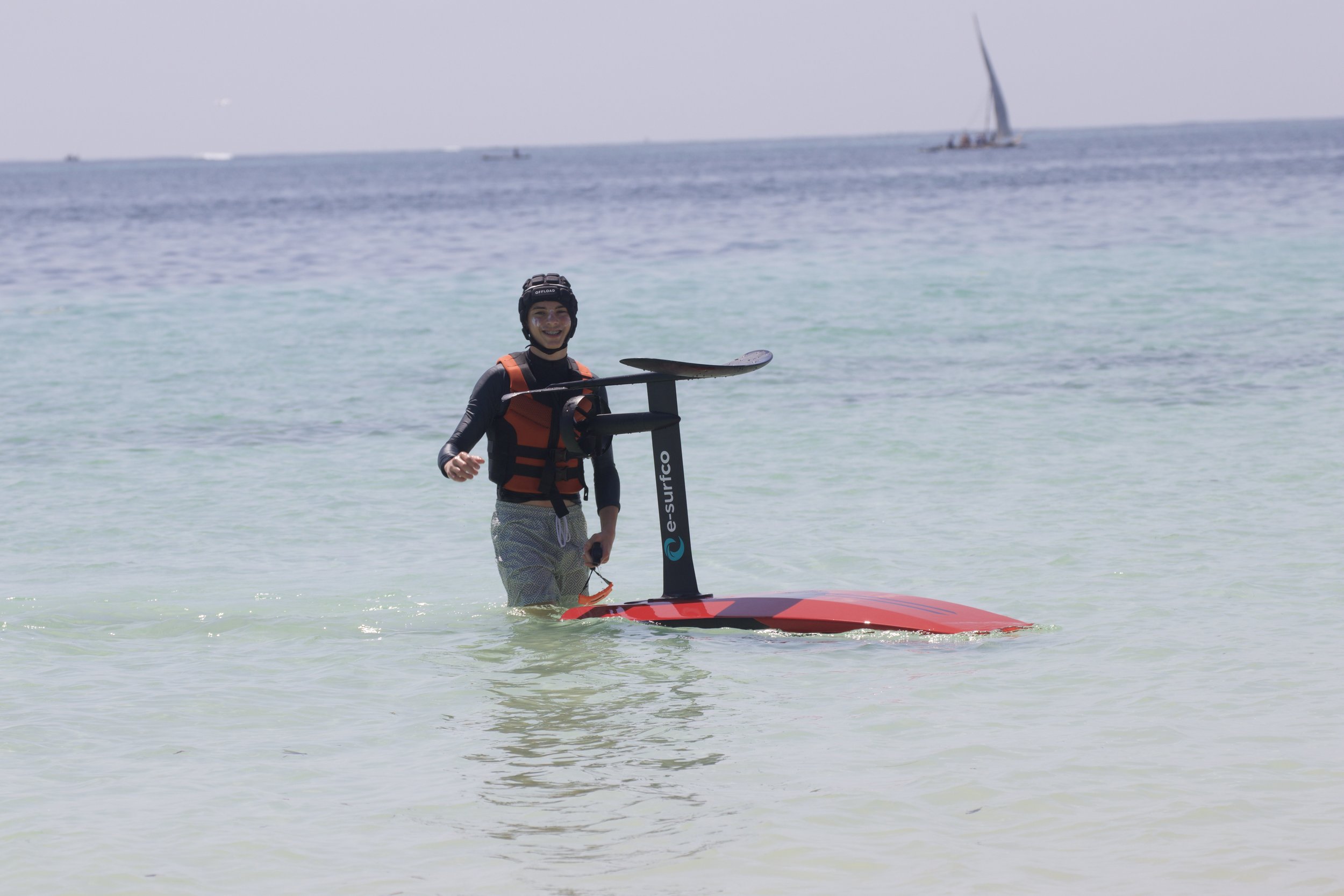 Person in a life jacket holding a stand-up paddleboard in shallow water at the beach, with sailboats and the ocean in the background.