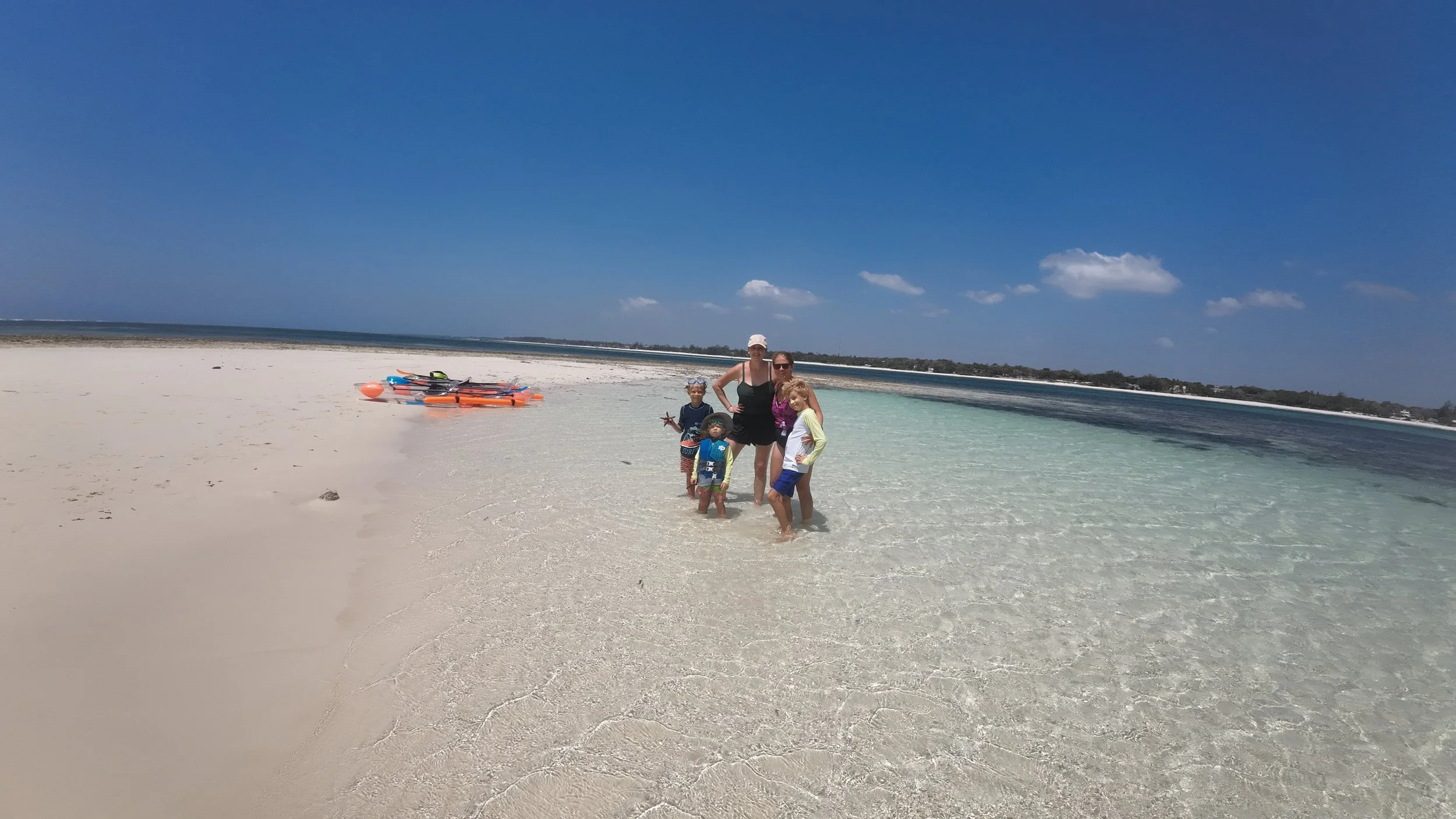 A woman and four children standing in shallow clear water at the beach, with kayaks on sand nearby and a clear blue sky overhead.