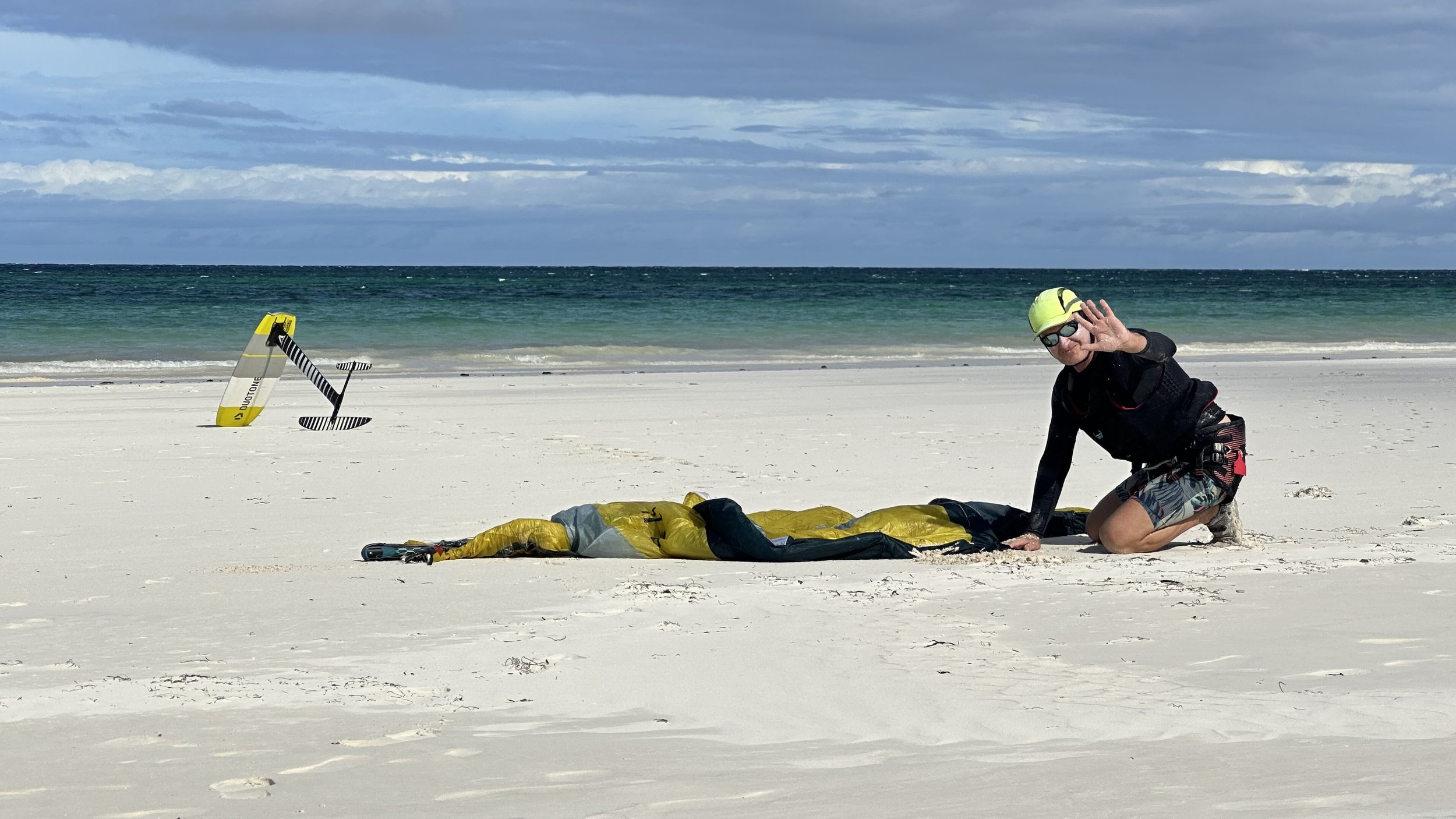 Person kneeling on sandy beach with wings laid out, preparing for kiteboarding, with water and cloudy sky in the background.