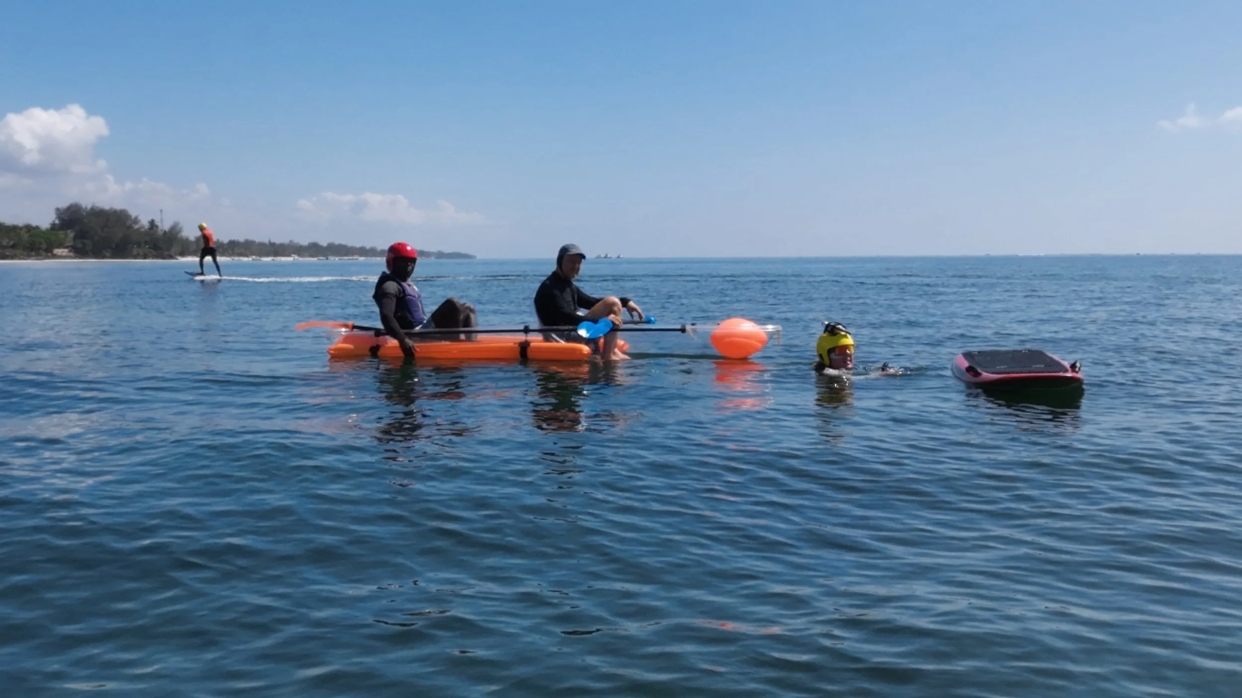 Two people sitting on an orange paddleboard with a buoy, a person in yellow helmet swimming nearby, and a person standing on a paddleboard in the background on calm ocean water under a clear blue sky.