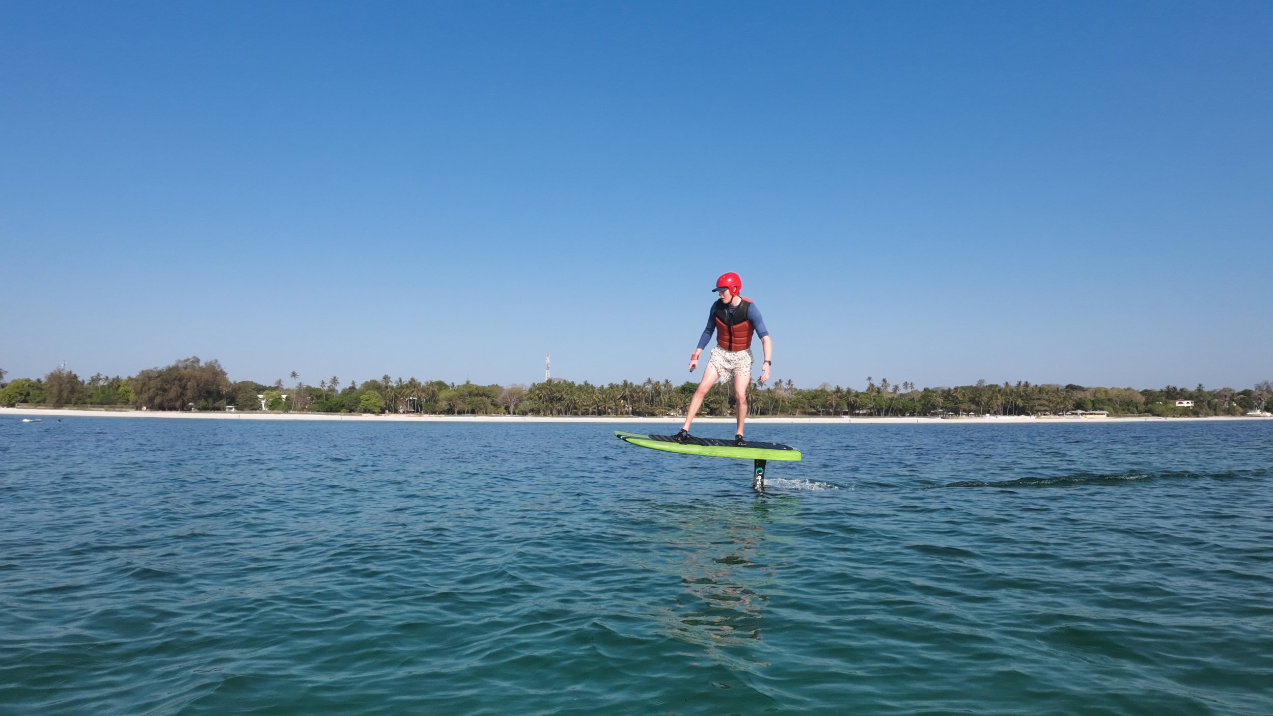 Person riding a hydrofoil in the water under a clear blue sky with a distant shoreline and trees.