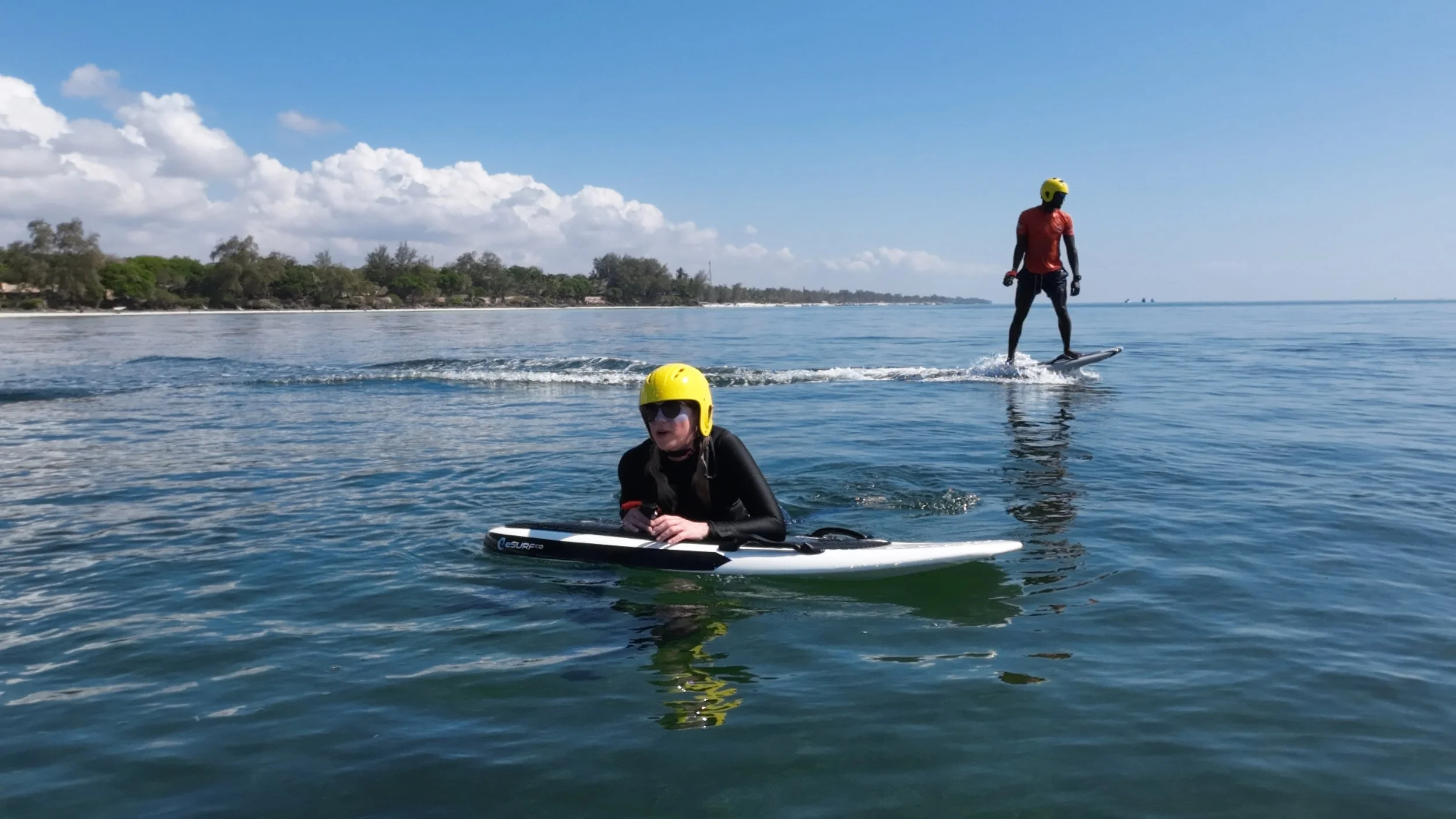 Two people stand-up paddleboarding on calm water under a partly cloudy sky, with a distant shoreline visible.