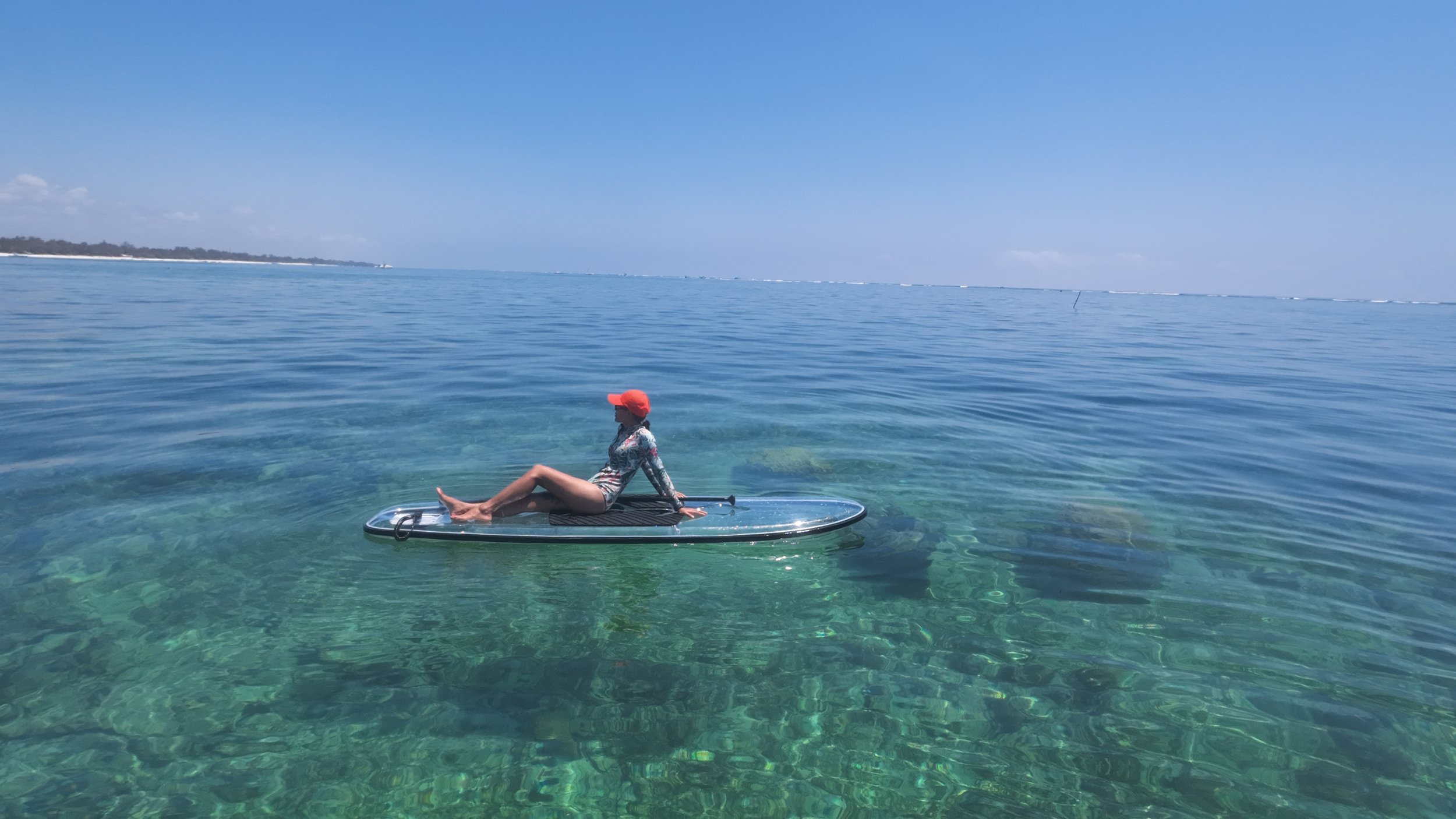 A woman sitting on a paddleboard in clear, blue ocean water with a distant shoreline and a bright blue sky.