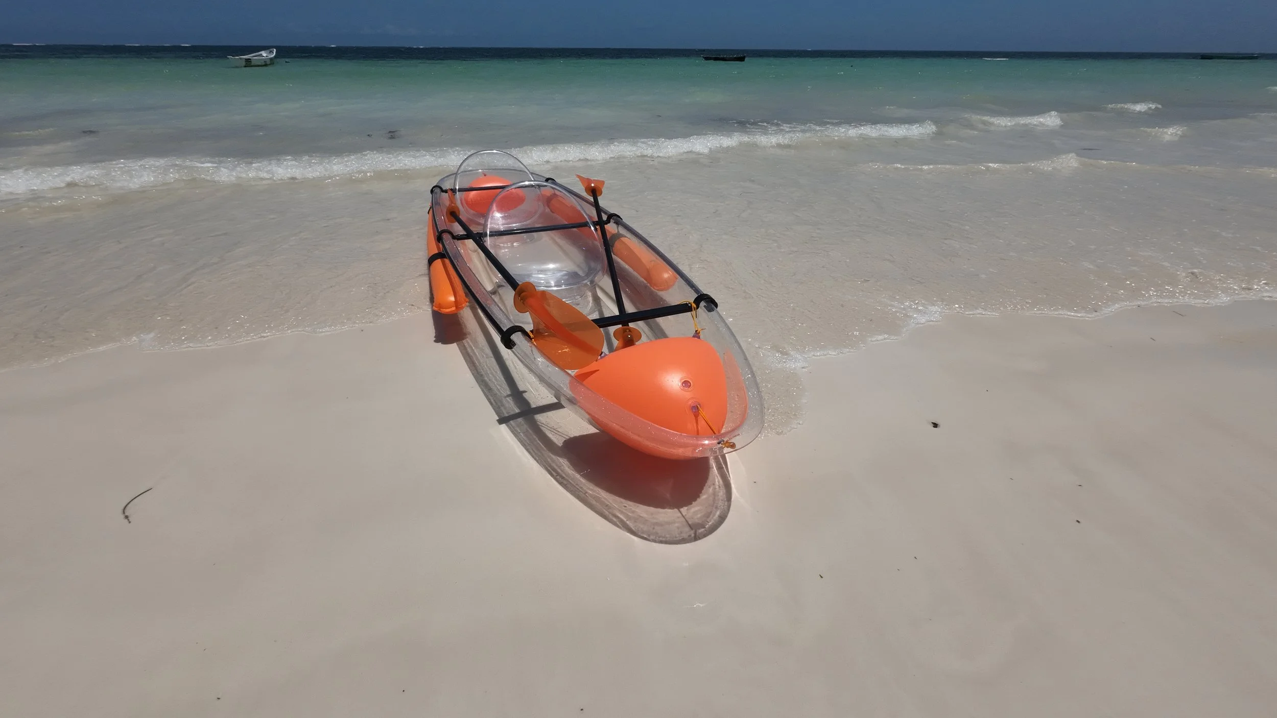 A clear kayak equipped with orange paddles, a large orange buoy, and a transparent canopy, resting on white sandy beach with gentle waves and a boat in the water in the background.
