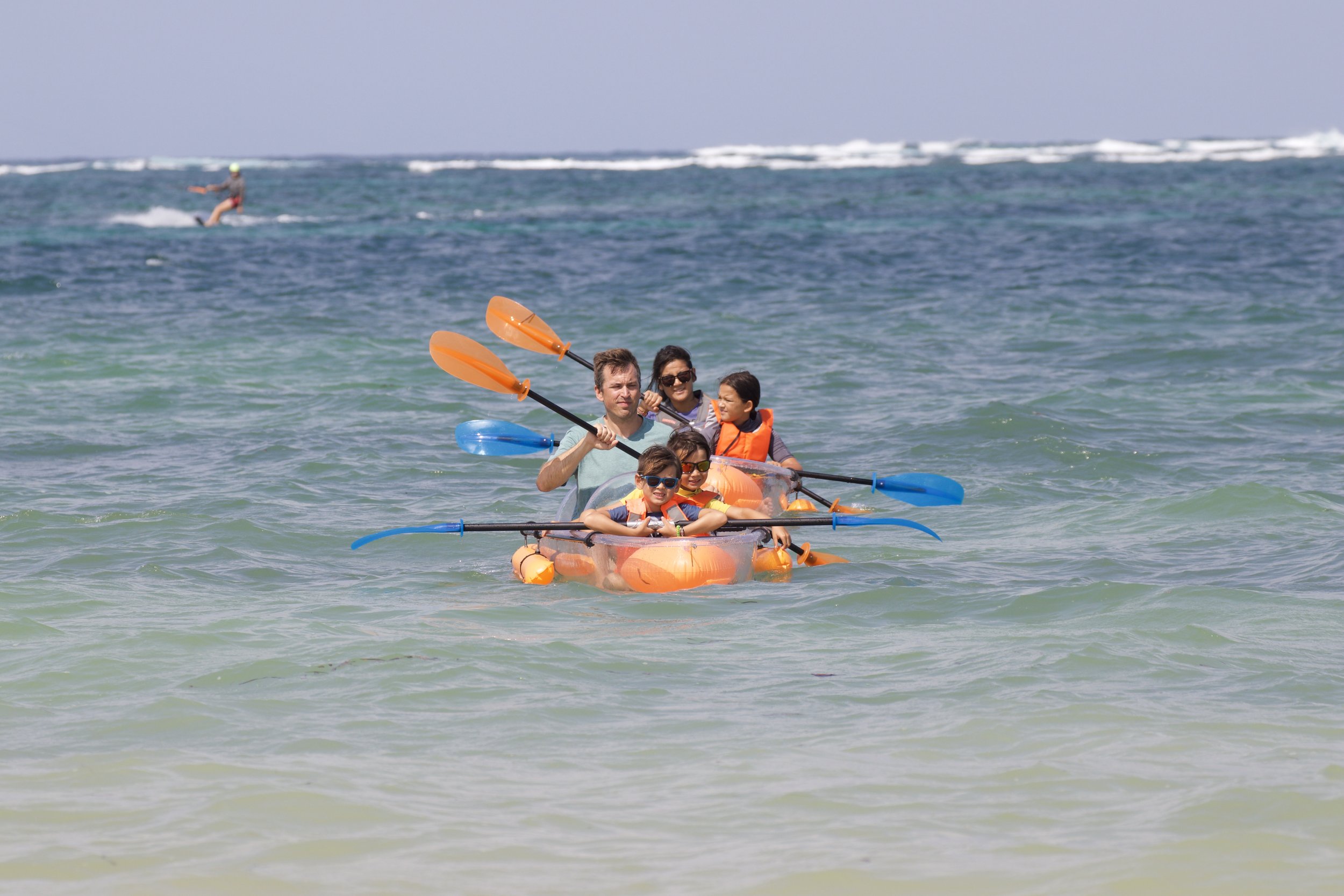 A family of five in a small orange kayak paddling in the ocean, with a person in the background wakeboarding.