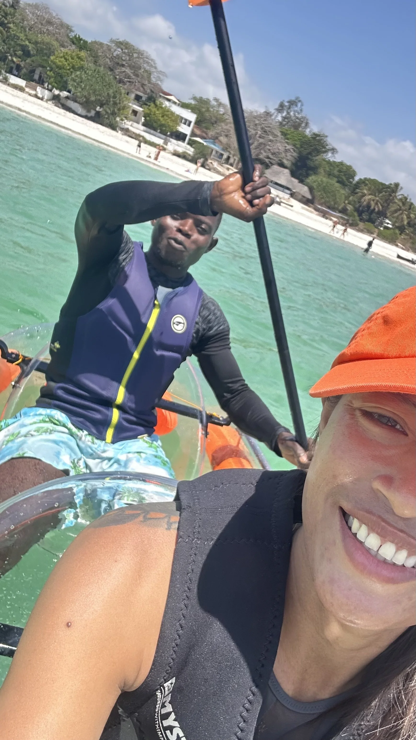 Two people taking a selfie on a boat in a tropical location, with a shoreline, trees, and buildings in the background, and clear green water.